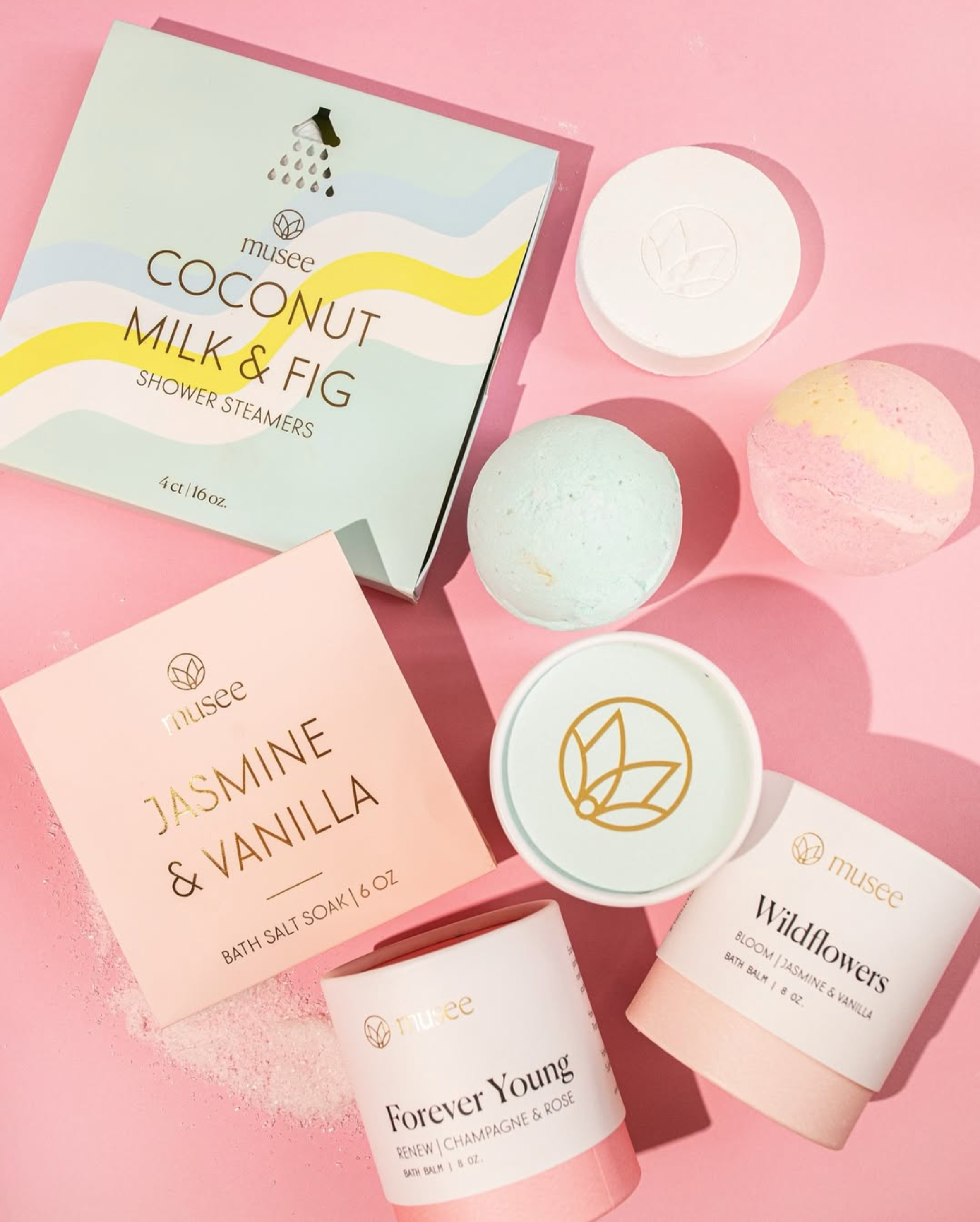A collection of bath and shower products, including a box of coconut milk and fig shower steamers, a bath salt box labeled jasmine and vanilla, a bath balm called Wildflowers, and a bath balm named Forever Young. The items are arranged on a pink back