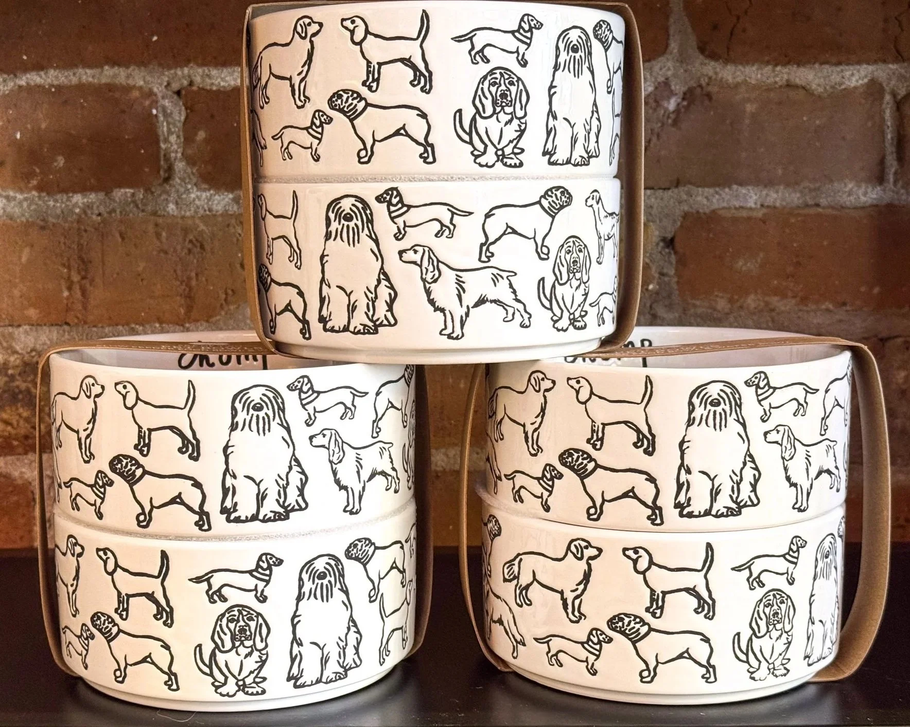 Three white ceramic bowls with black line drawings of different dog breeds on them, stacked in a pyramid formation against a brick wall background.