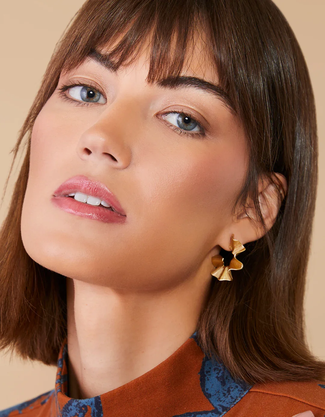 Close-up of a woman with shoulder-length brown hair, blue eyes, wearing gold flower-shaped earrings and a patterned top against a neutral background.