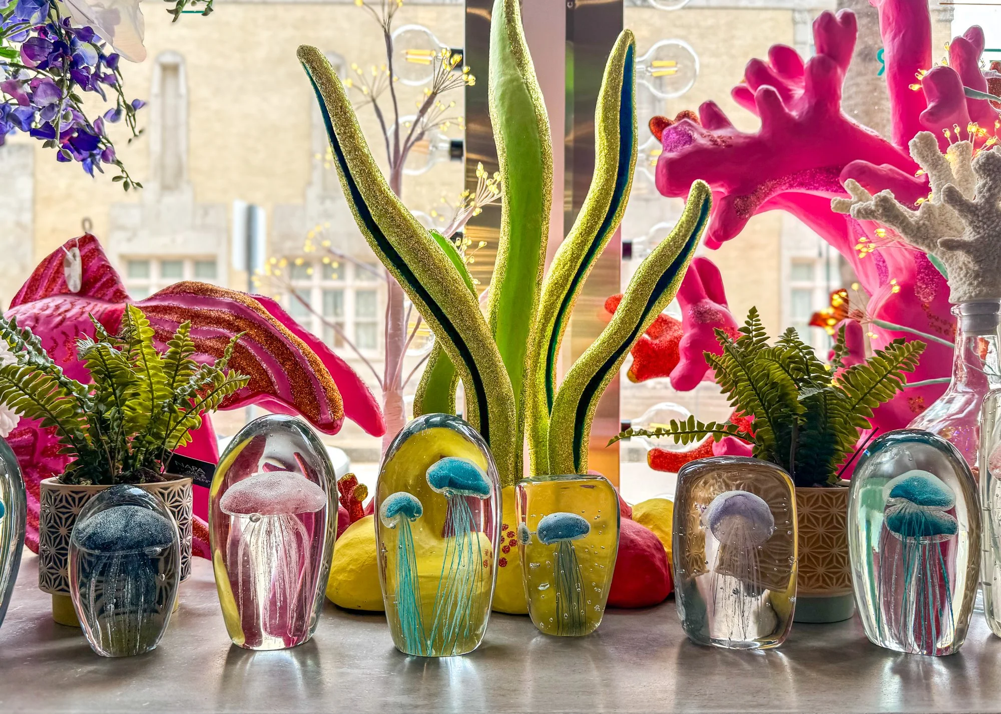 Colorful glass sculptures of jellyfish, pink anthuriums, green ferns, and decorative plants on a windowsill with a cityscape view in the background.