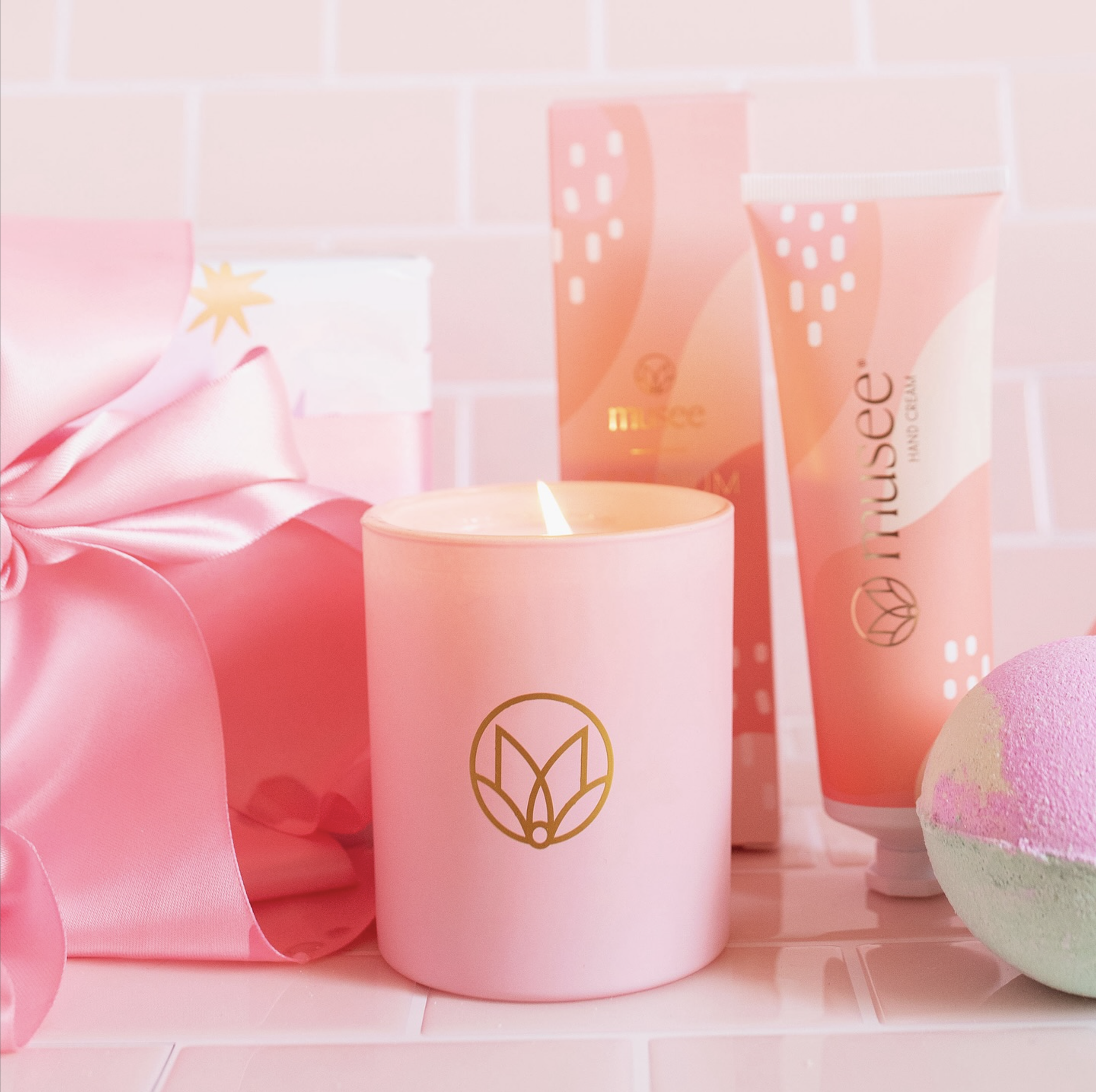 A pink candle with a gold geometric leaf logo burns next to a wrapped gift with pink ribbon, pink packaging, pink hand cream, and a pink and white bath bomb, all on a pink tiled surface.
