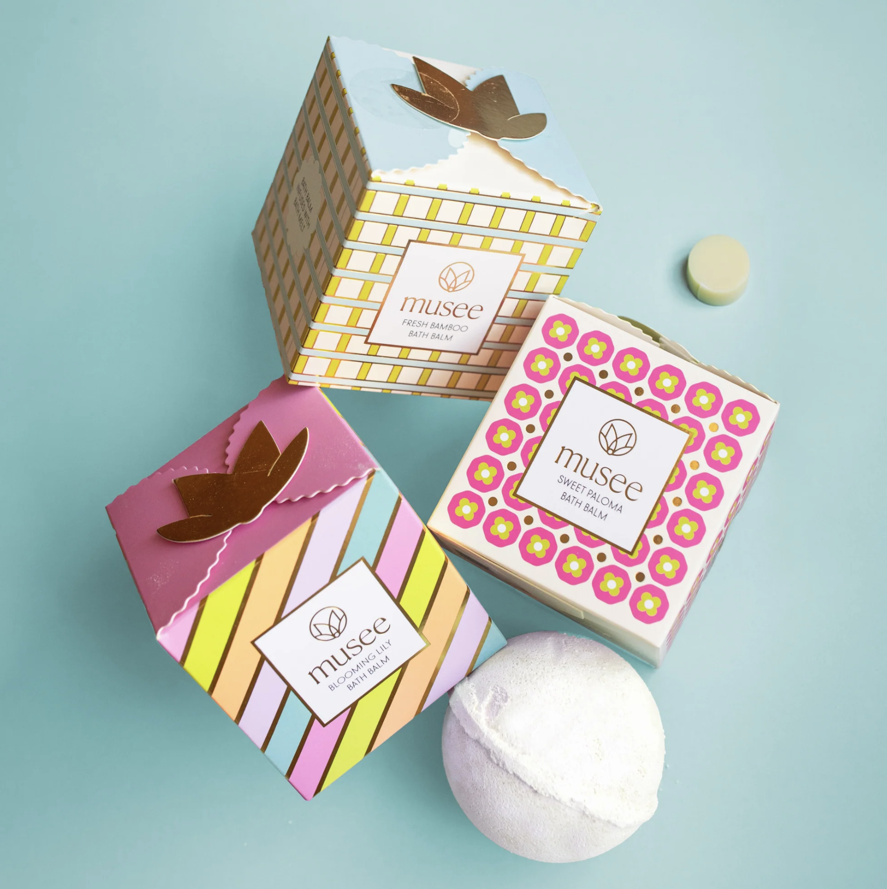 Three colorful boxed bath balms with floral and geometric patterns, a white bath bomb, and a small green round item on a light blue background.