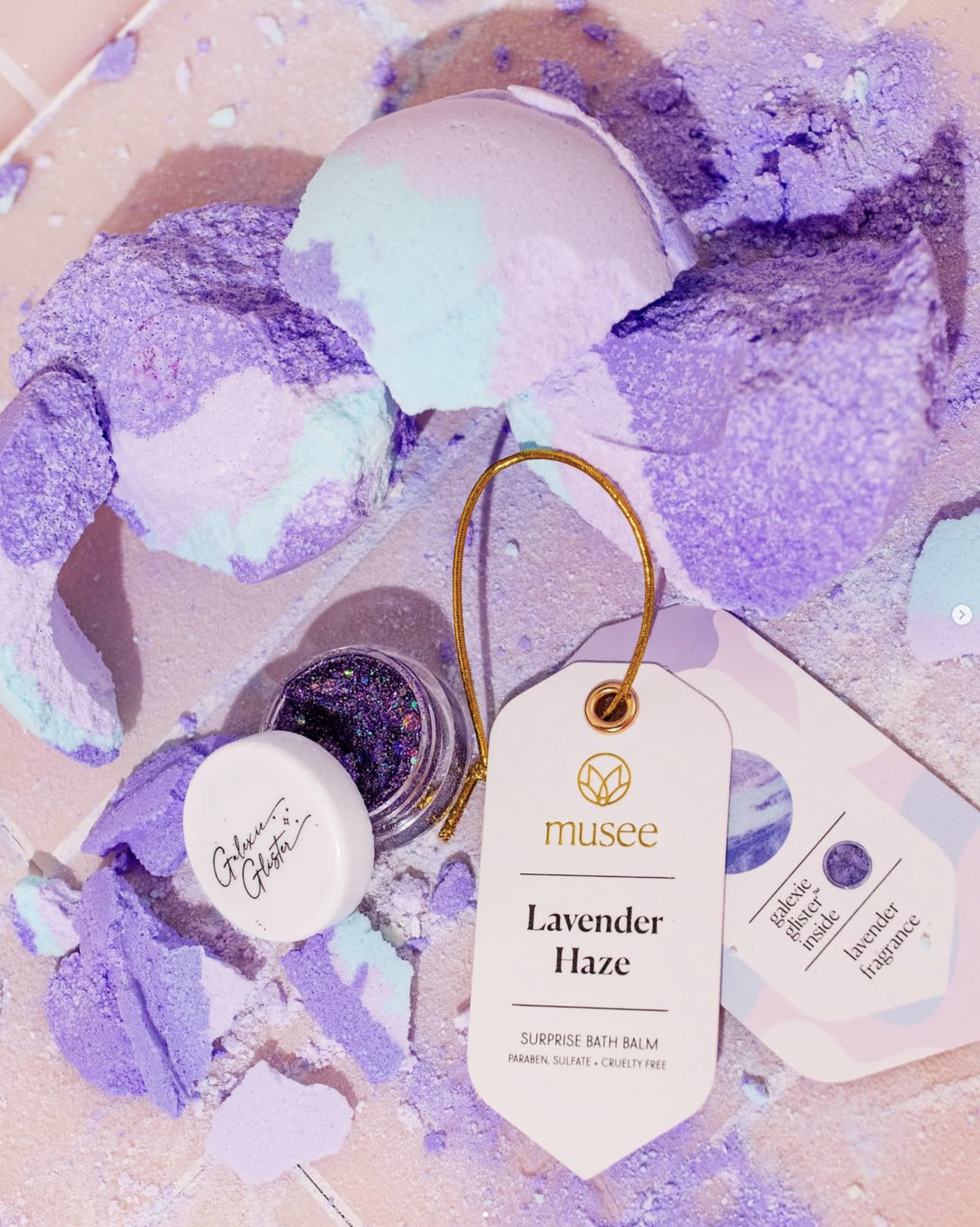 A bath bomb with purple and white colors, a small container of glitter, and a tag reading 'Lavender Haze' from Musee. The background features pastel-colored bath bombs or crushed bath bomb pieces in purple, pink, and blue.