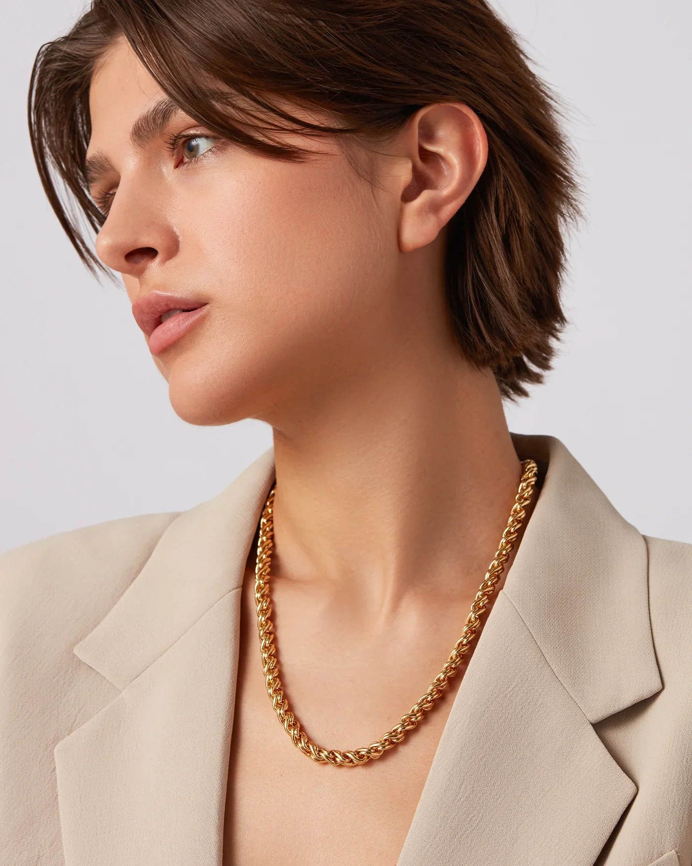 A woman with short brown hair wearing a beige blazer and a gold chunky necklace.