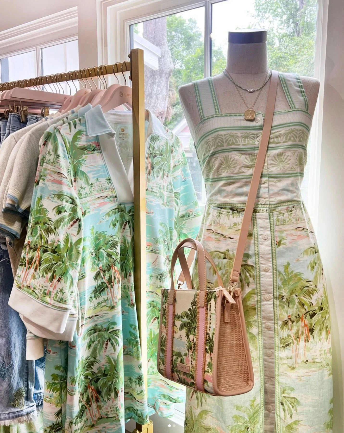 Display of tropical-themed clothing including a dress on a mannequin, a matched shoulder bag, and a shirt on a clothing rack, all featuring palm trees and scenic prints, inside a retail store with a window in the background.