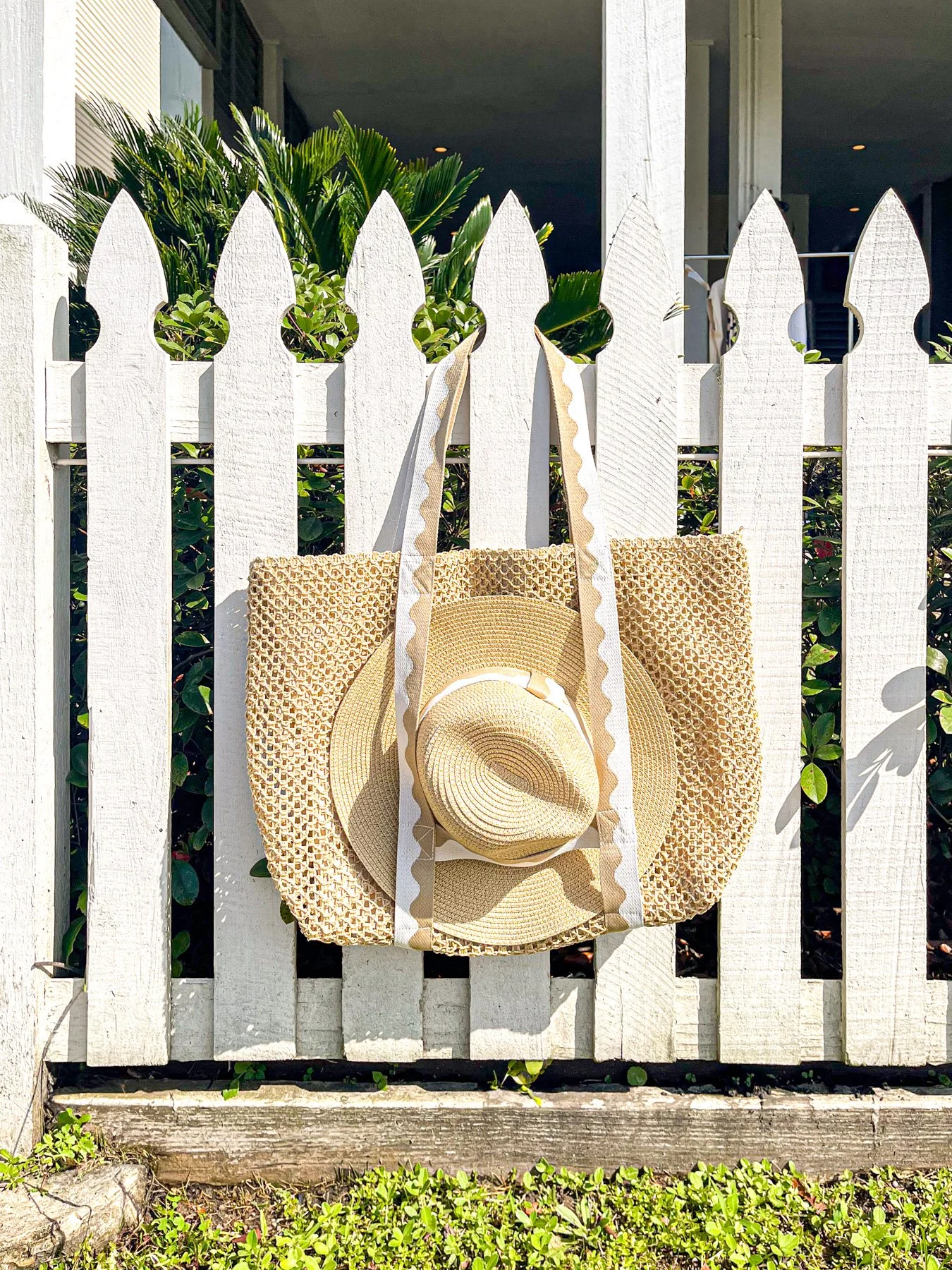 A beige sunhat hanging on a white wooden fence with green plants behind it.