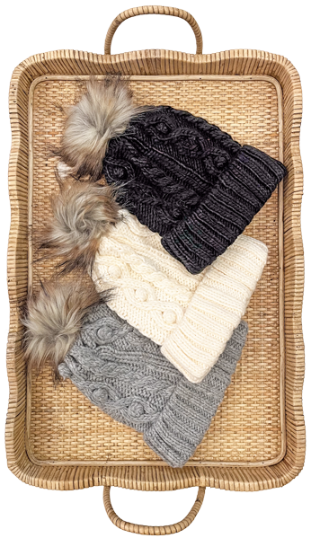 A wicker basket containing three knitted winter hats with pom-poms, in black, cream, and gray colors.