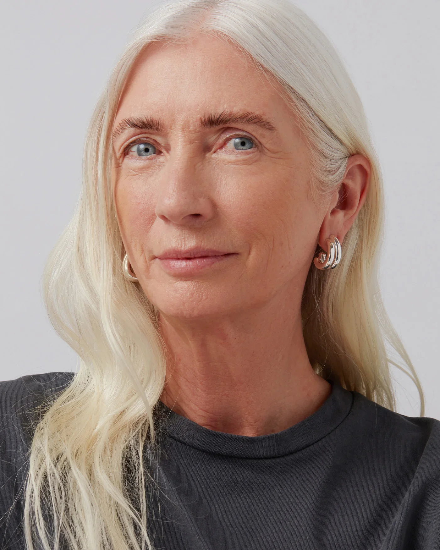 Close-up portrait of a middle-aged woman with long blonde hair, blue eyes, wearing a dark top and silver hoop earrings, against a plain white background.