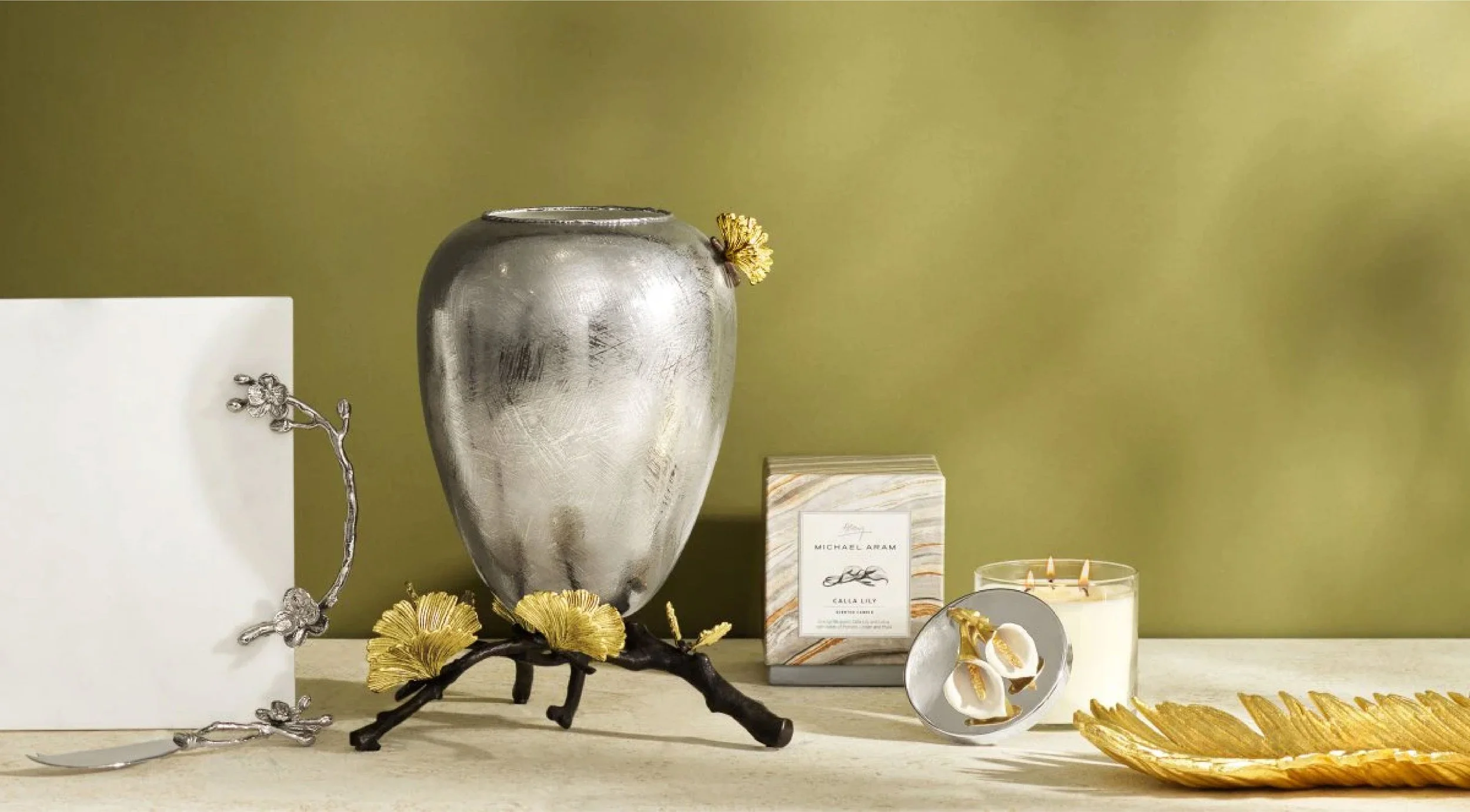 Decorative home items including a silver vase with gold accents, a candle in a glass holder, a gold leaf platter, and a silver and white tray with floral embellishments, displayed on a surface against a light green background.