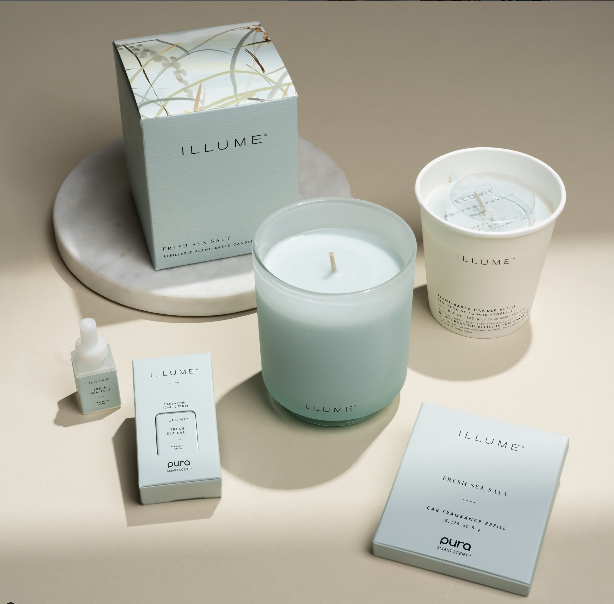 Illume Fresh Sea Salt candle and fragrance products displayed on a beige background, including a boxed candle, a glass candle, a cream-colored holder, a fragrance oil bottle, and a car fragrance refill package.