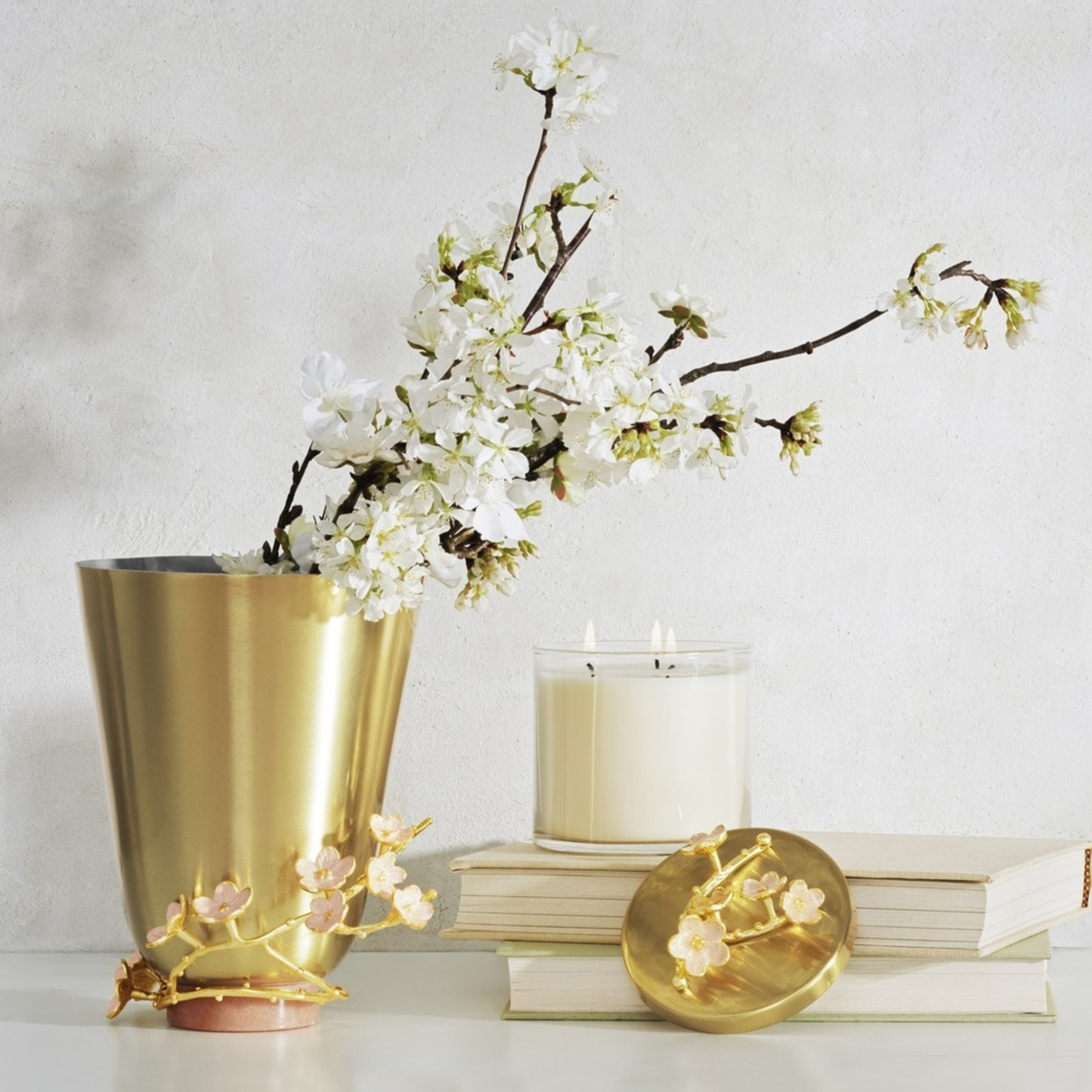 A gold vase with white flowers and branches, a lit white candle, two stacked books, a gold circular jewelry dish with pink flower accents, and decorative pink and gold branch design.