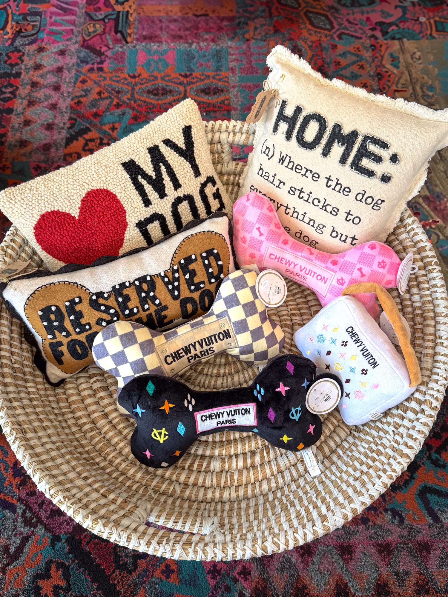 A collection of decorative pillows and plush toys in a wicker basket, with a colorful patterned carpet underneath. The pillows feature humorous and love-themed text, while the plush toys are shaped like bones with brand logos and colorful patterns.