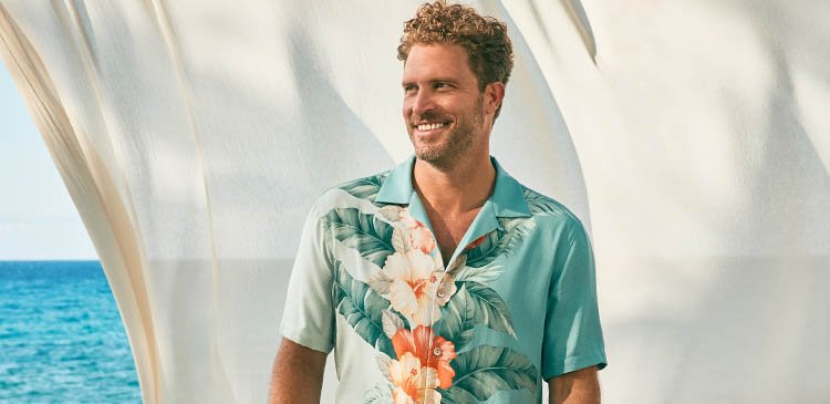 A man with curly hair and a beard wearing a tropical shirt, smiling, with a seashell and ocean in the background.