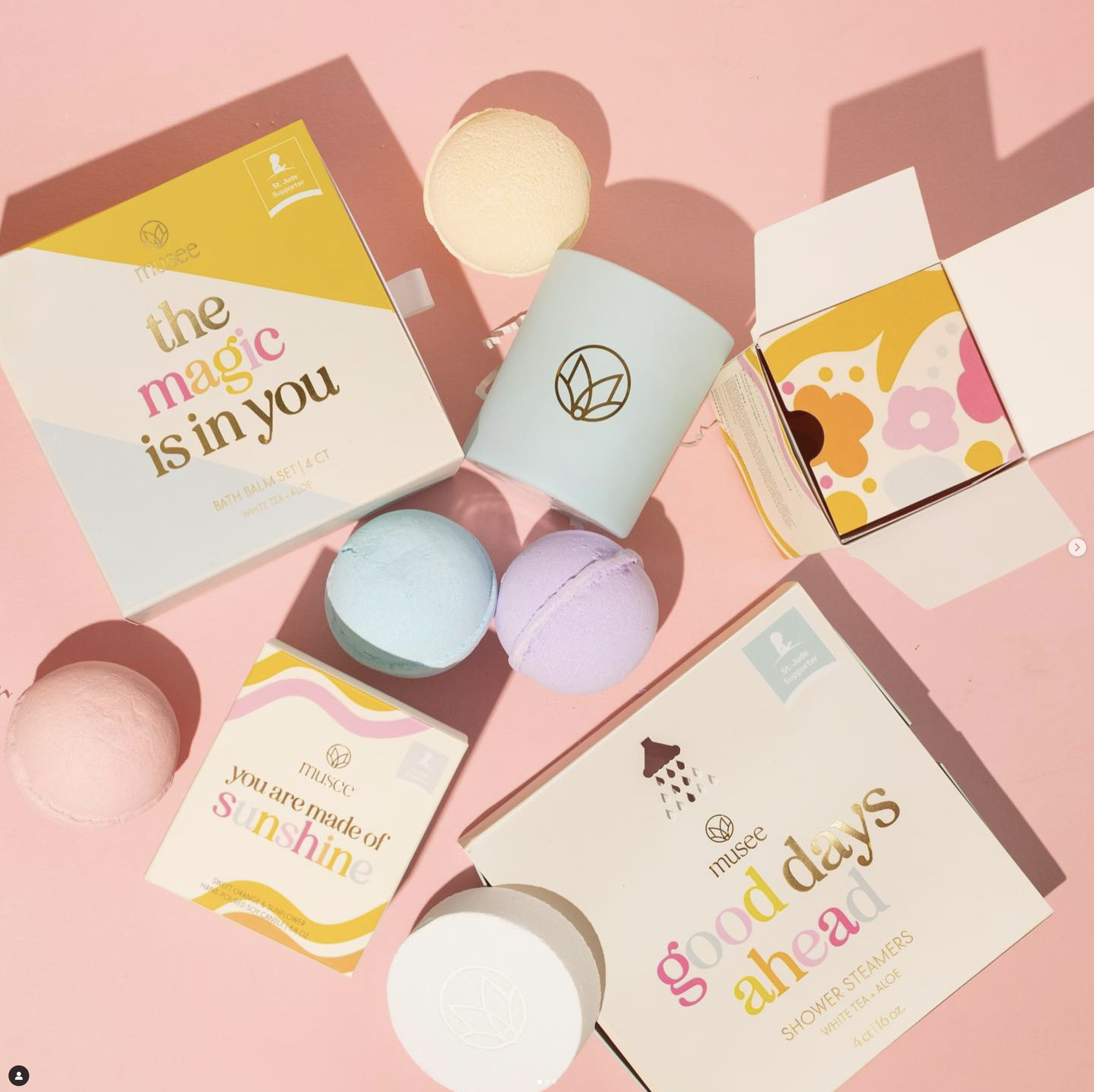 Various colorful bath products and packaging on a pink surface, including bath bombs, boxes with uplifting messages, and containers with leaves and floral designs.