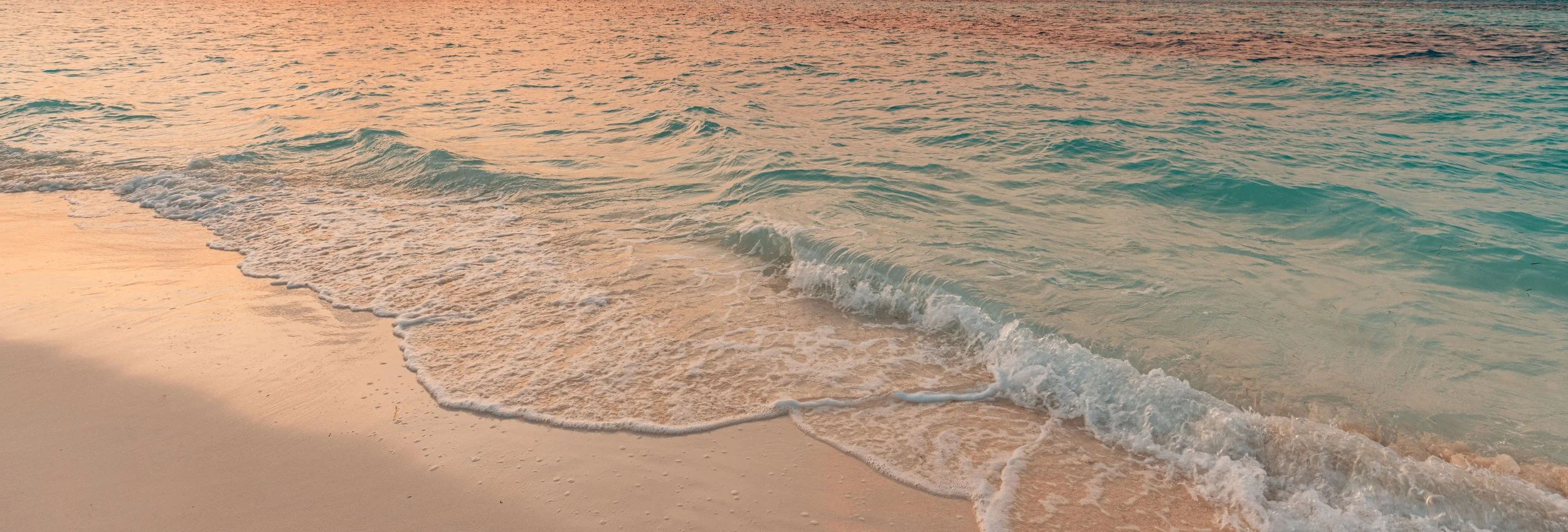 Wave gently crashing on a sandy beach at sunset with soft pink and orange hues in the sky.