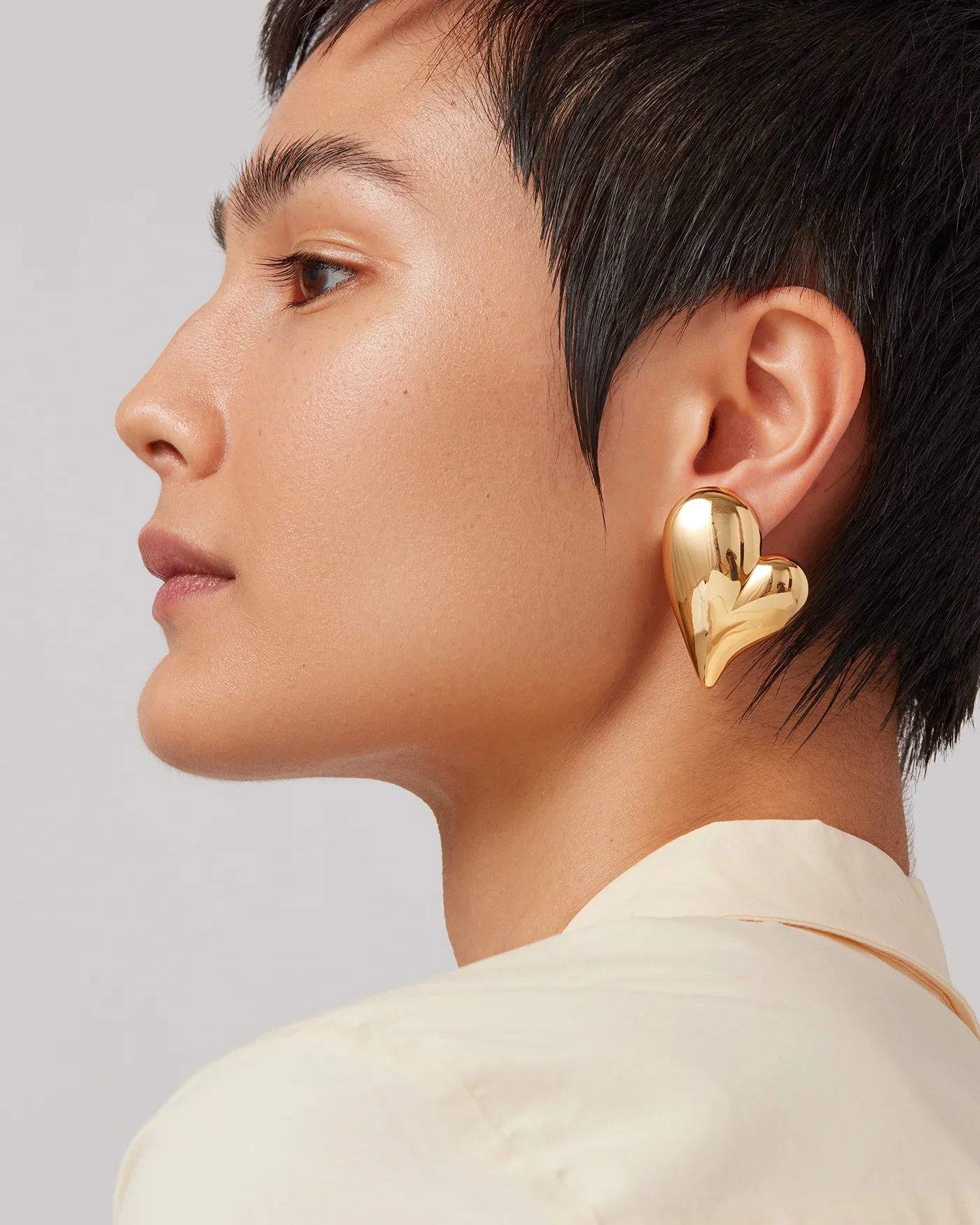A woman with short black hair wearing a large gold heart-shaped earring and a light-colored satin shirt.