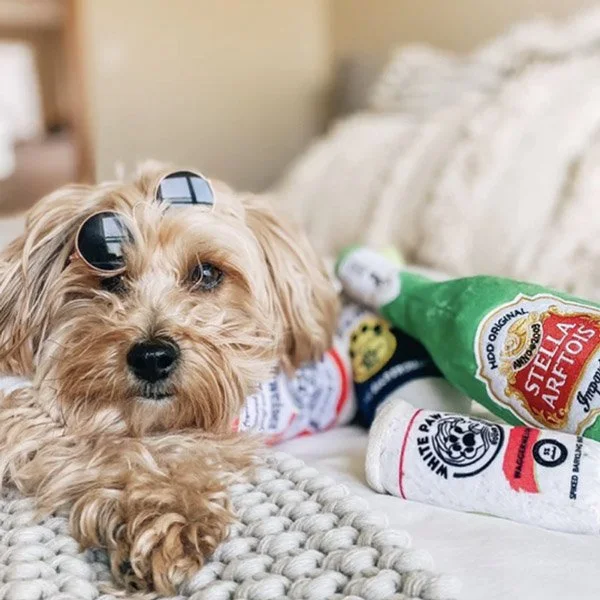 Cute small dog with sunglasses lying on a bed next to green and white hot sauce bottles.