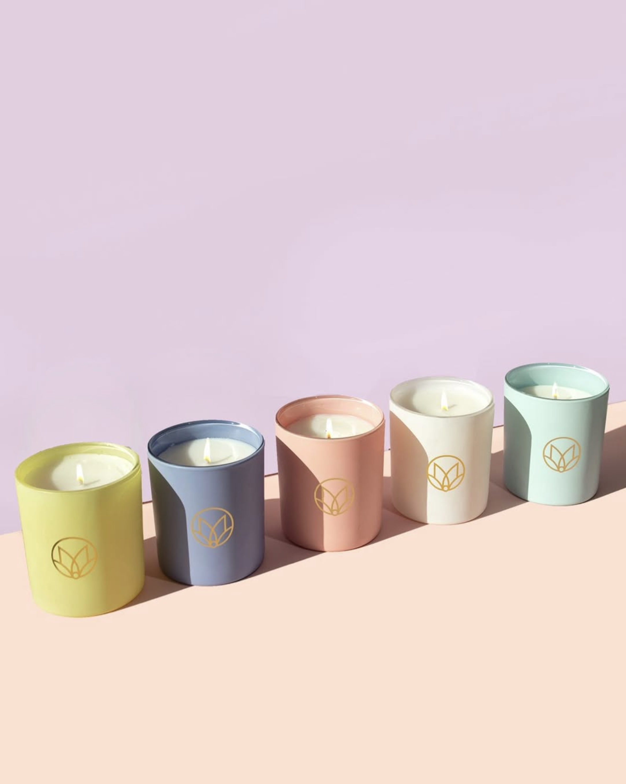 Five colorful candles in pastel-colored holders with a gold logo, arranged on a pink surface with a light purple background.