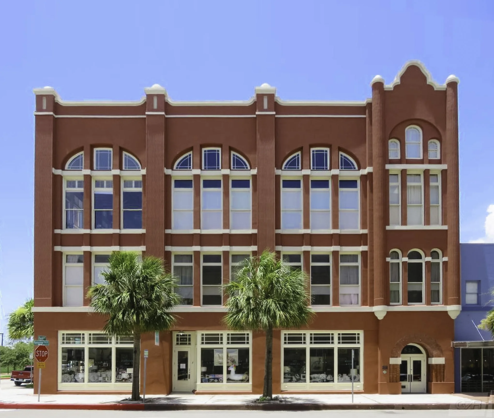 A red brick building with four floors, large arched windows, and decorative white accents. Two palm trees are in front, and part of a blue building is visible on the right. Clear blue sky in the background.