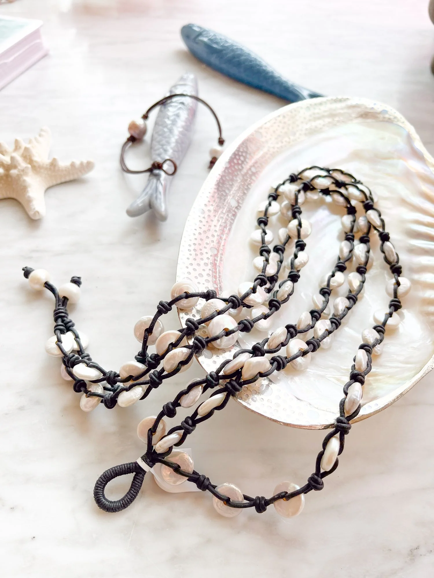 Beach-themed jewelry including a pearl necklace and shell necklace on a large shell plate, with starfish and seashells in the background.