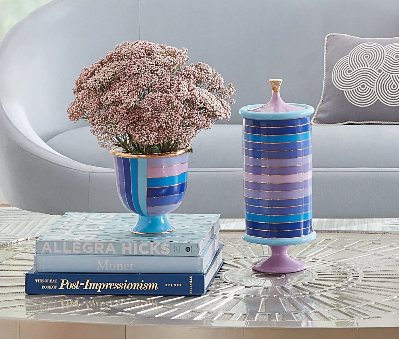 Modern living room setting with a striped ceramic vase holding pink flowers on a coffee table. A matching lidded container is next to a stack of books with titles visible, with a gray couch and decorative pillow in the background.