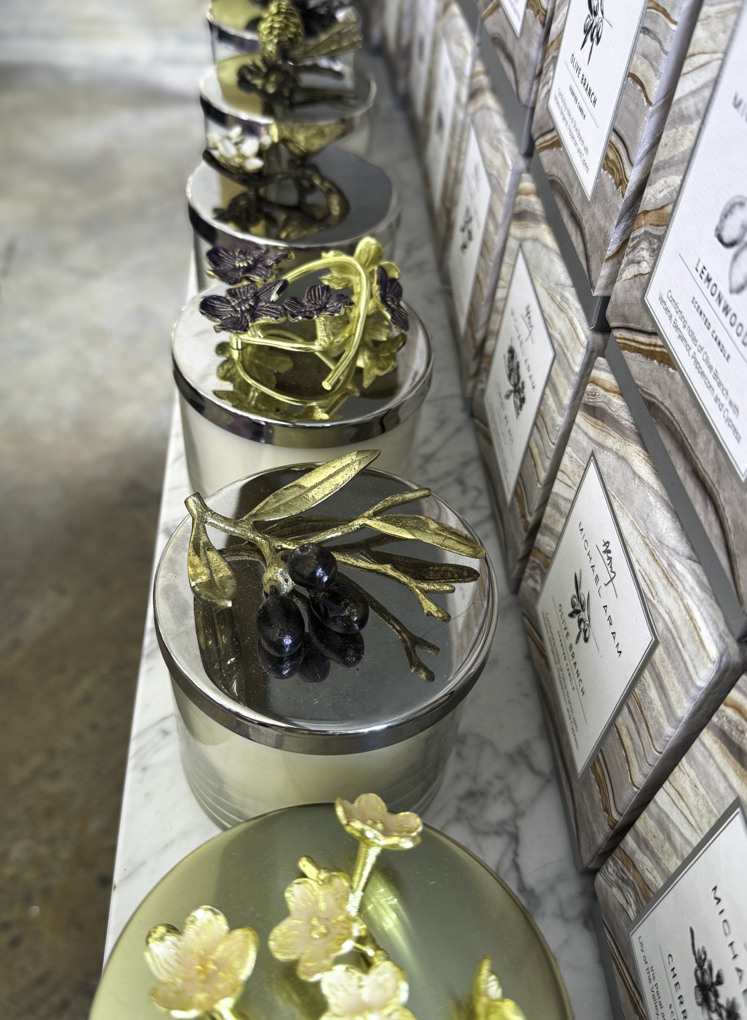 Decorative candle jars with gold floral and olive branch designs, alongside boxed scented candles on a marble surface.
