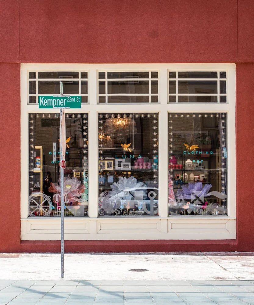 The storefront window of Luna, a boutique shop, on a corner with brick walls, decorated with lights and flowers. The window displays various items, including clothing, pet accessories, and gifts. A sign on the window lists categories like books, clothing, pet, and gifts. There is a street sign indicating the corner of Kempner 22nd Street, and the sidewalk in front of the store is visible.