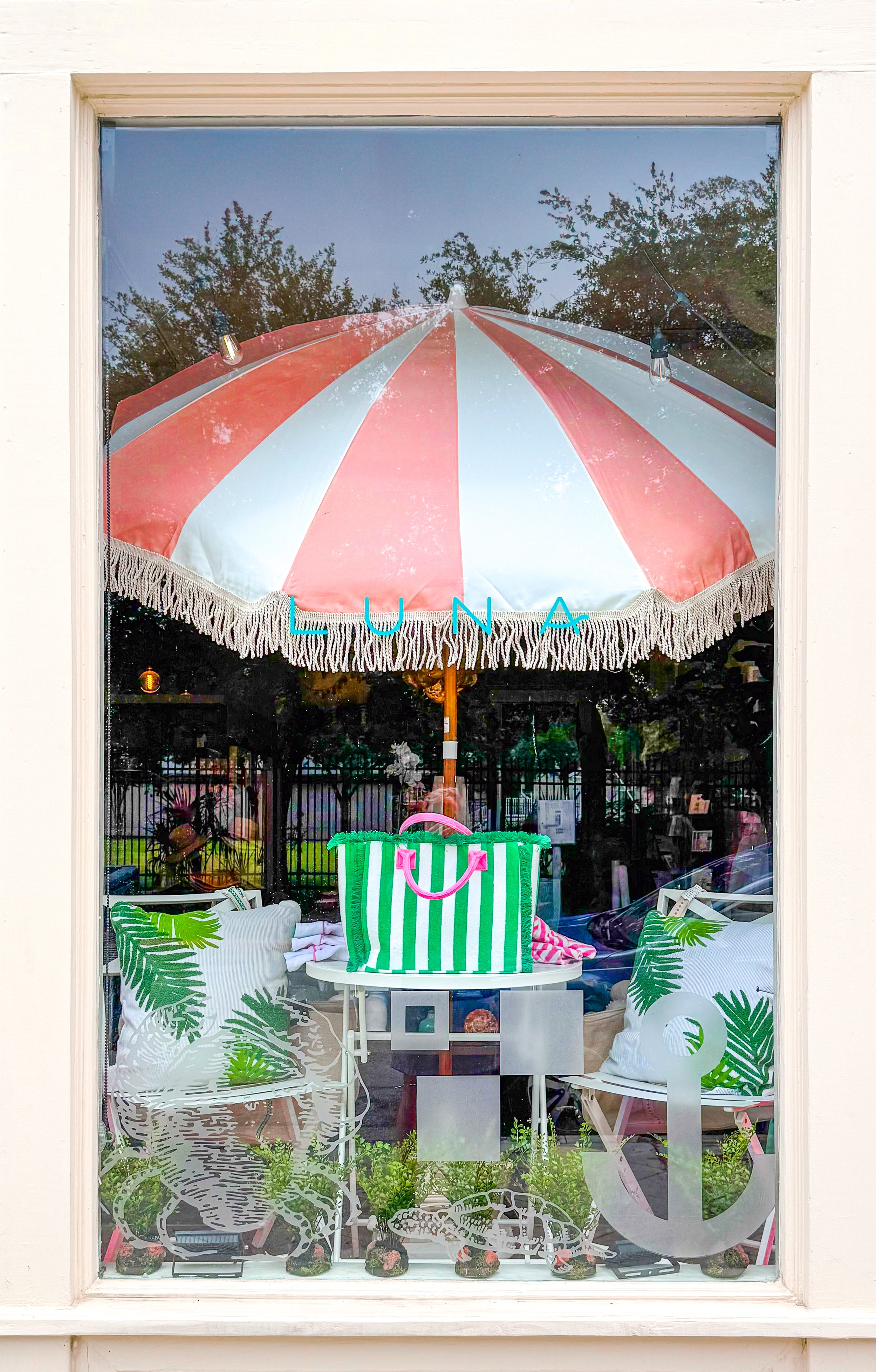 A shop window display with a pink and white striped umbrella, green and white striped bag, cushions with tropical leaf prints, and nautical-themed decor.