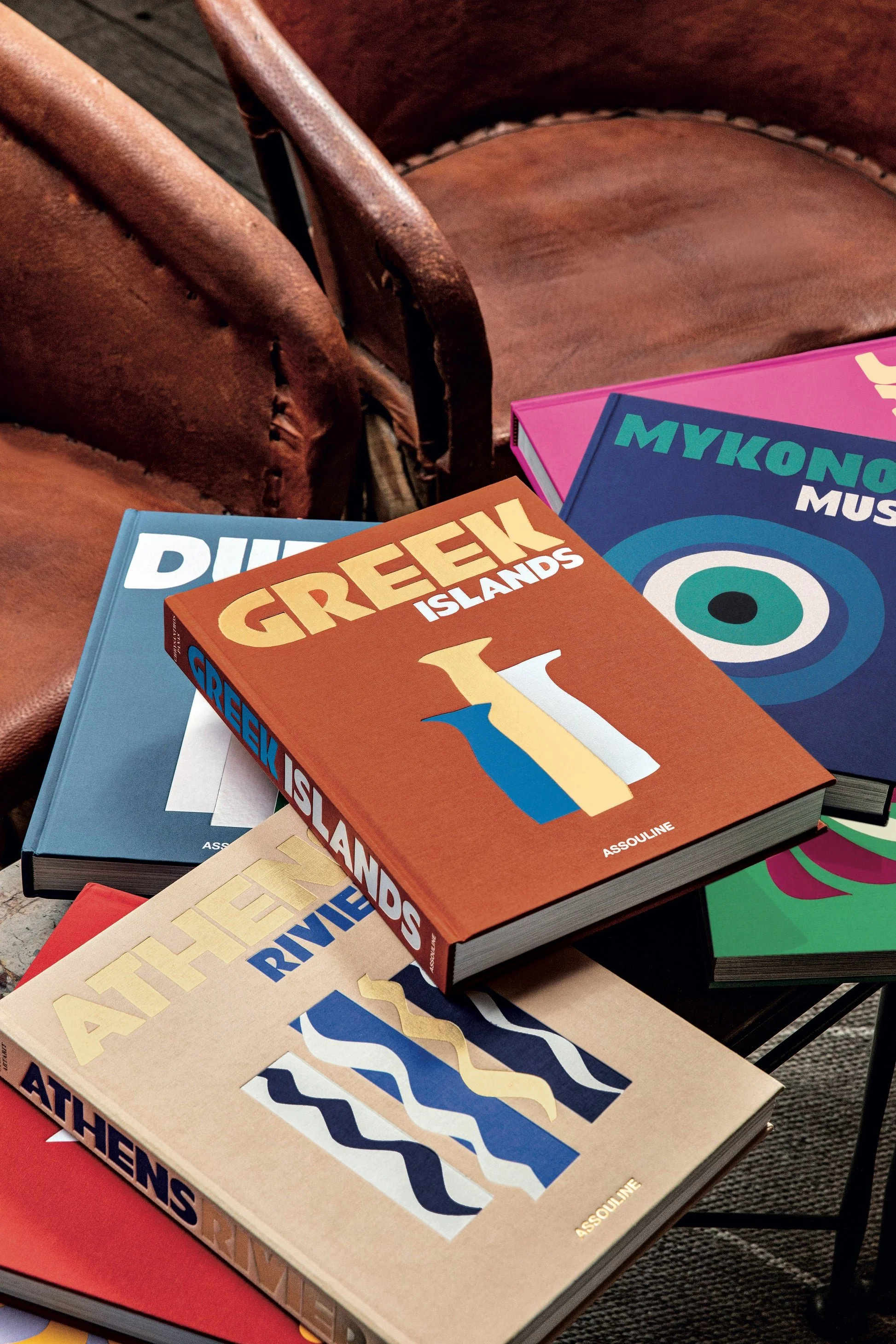 Assortment of colorful travel books on Greek destinations on a table with brown chairs.