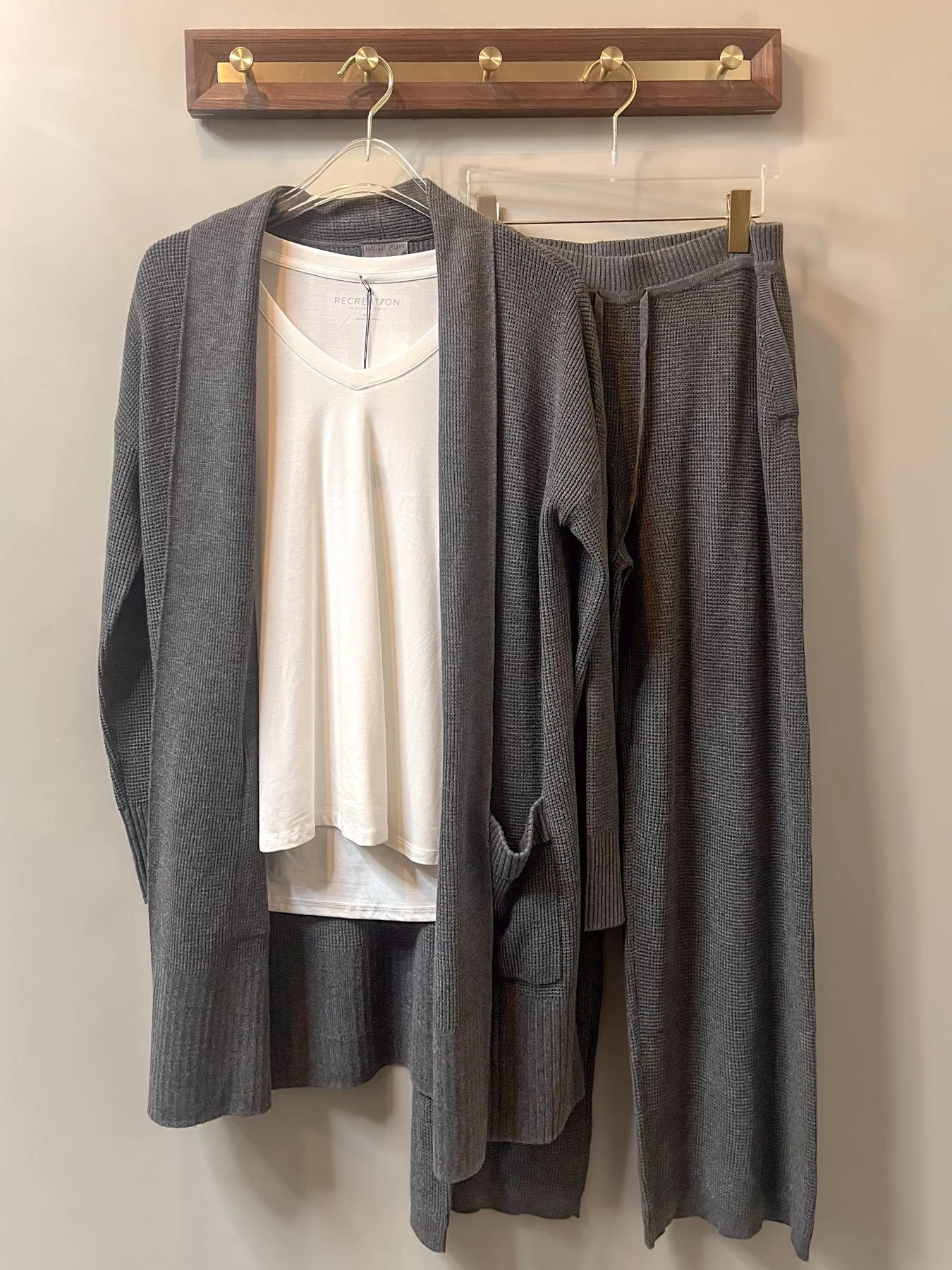Gray cardigan, gray pants, and a white t-shirt hanging on hooks and a wooden rack.