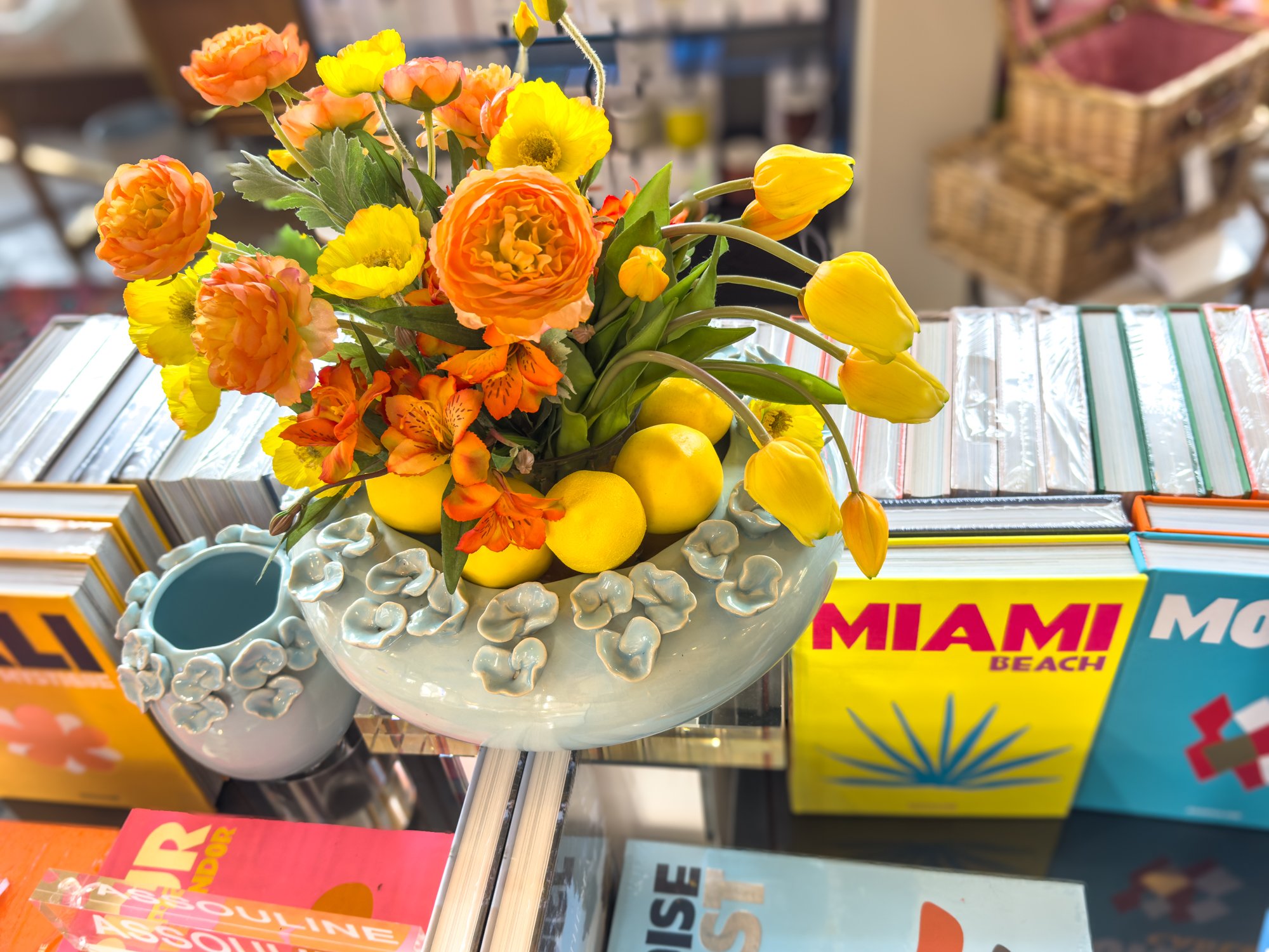 A floral arrangement with orange and yellow flowers in a white vase decorated with small white dog figurines, placed on a glass surface with travel guides around it.