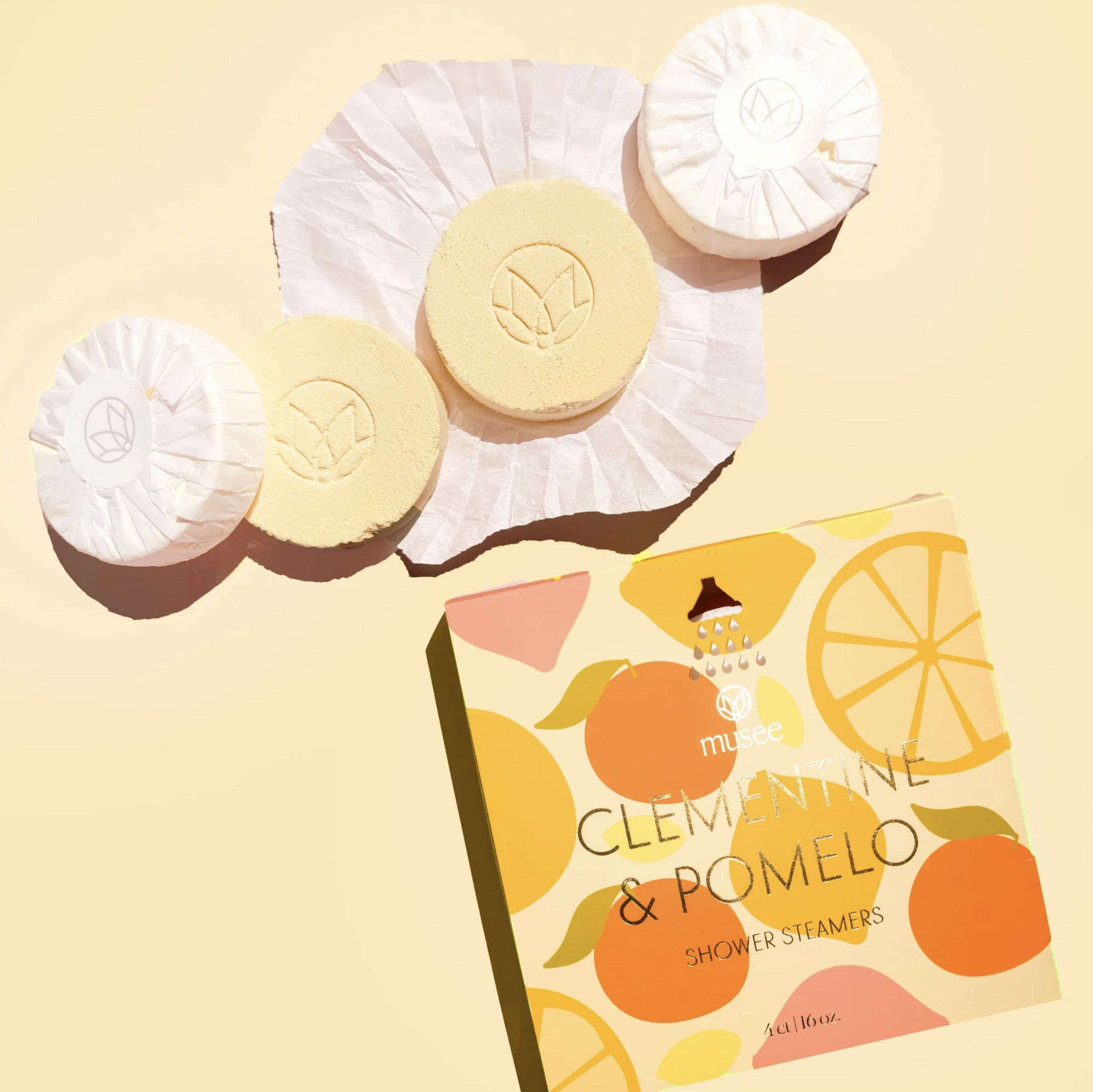 Three round, pastel yellow bath bombs with embossed citrus fruit logos, partly unwrapped, placed on white tissue paper, with a colorful box labeled "Clementine & Pomelo Shower Steamers" featuring citrus illustrations.