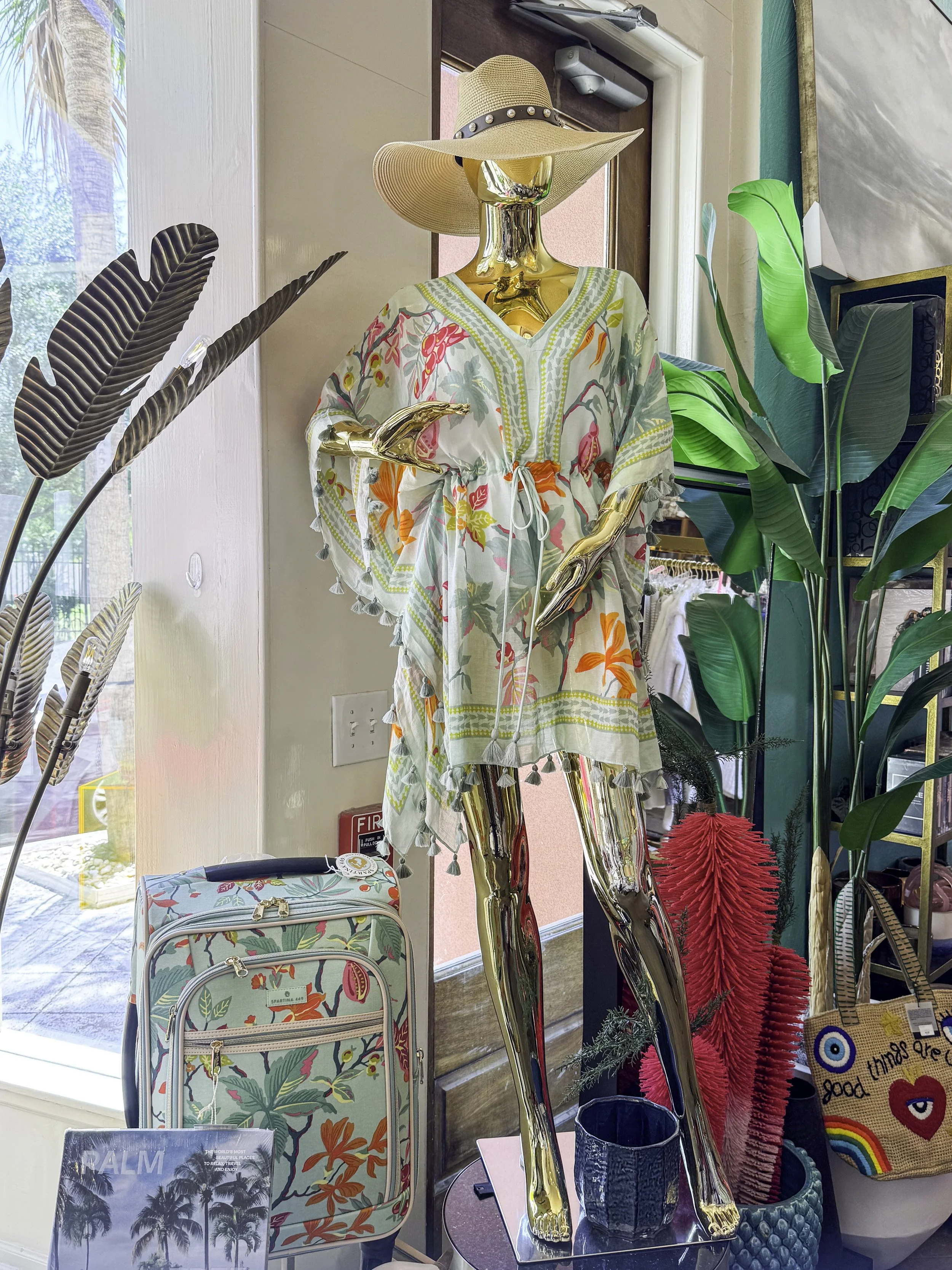 Mannequin dressed in a floral kaftan and a wide-brimmed hat, displayed in a store with plants and a floral-patterned suitcase.