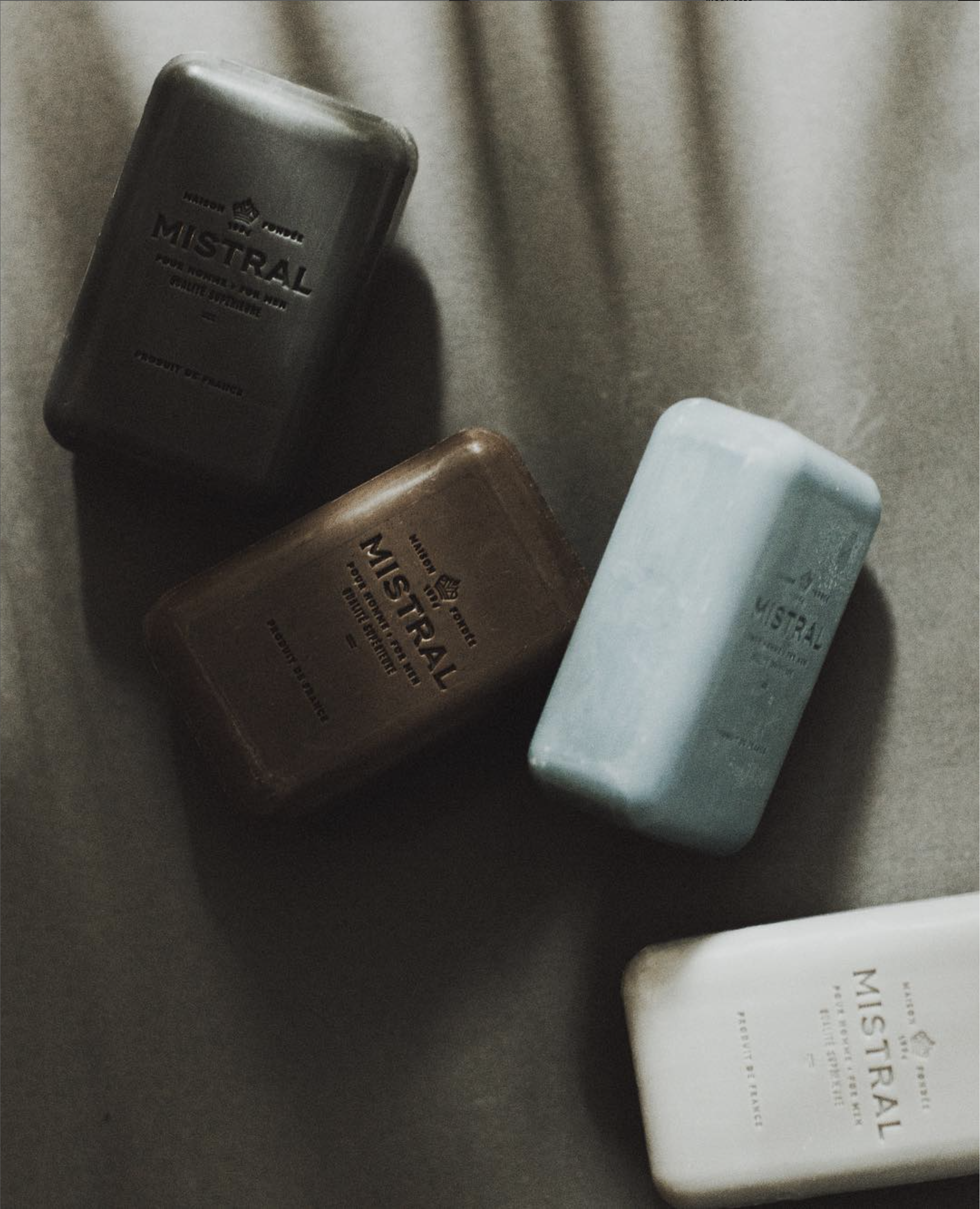 Four bars of Mistral soap in different colors on a gray surface.