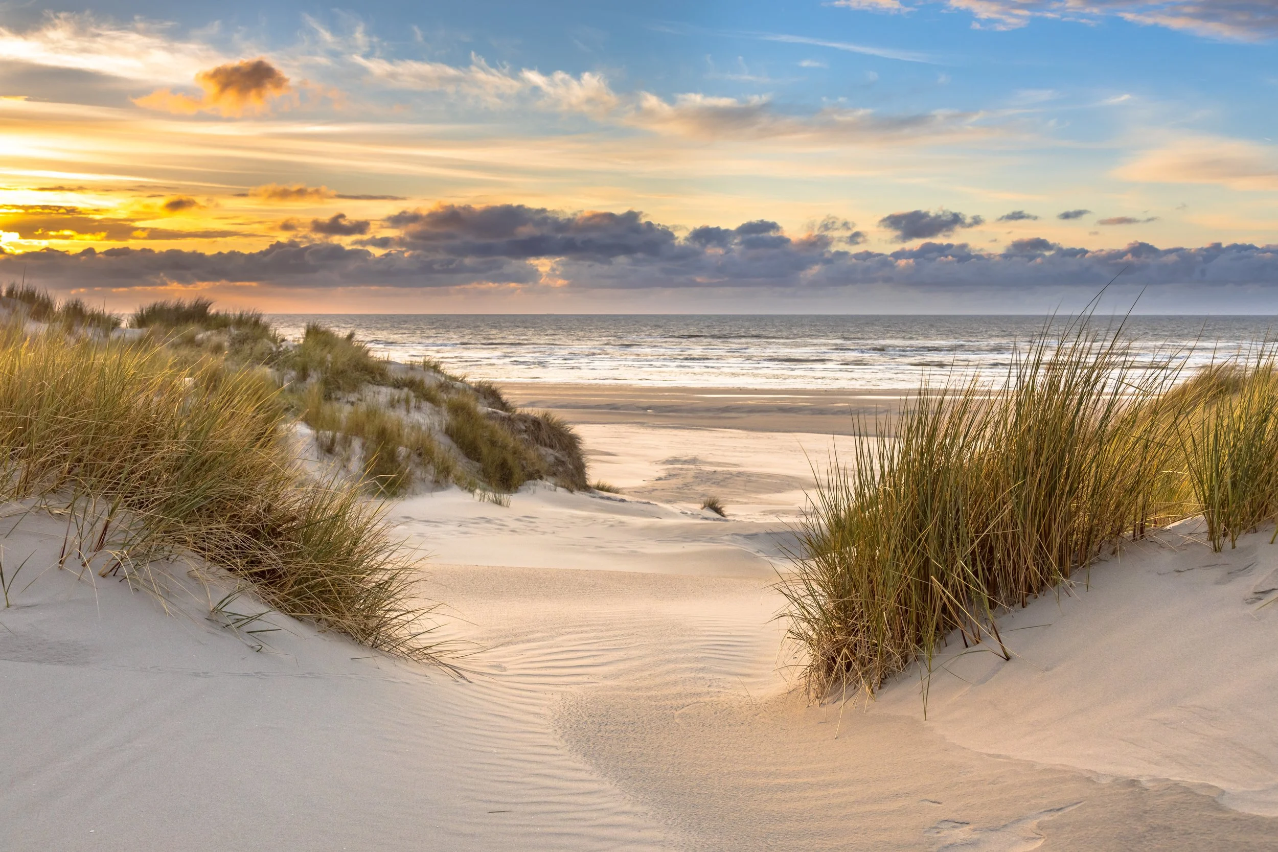 Sunset over the beach with sand dunes and tall grass, calm ocean waves, and a partly cloudy sky.