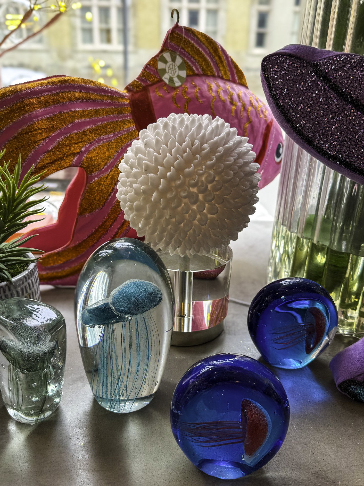 Decorative display with glass jellyfish paperweights and vibrant ornaments, including a pink fish sculpture with gold accents and a spherical white textured object, set on a window ledge.