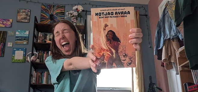 picture of a brunette girl with a joyous expression holding up a book titled "Aarya Dalton Didn't Sign Up For This"