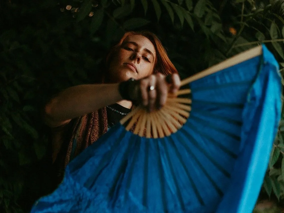 A woman with red hair holding a blue paper fan, surrounded by dark green foliage.