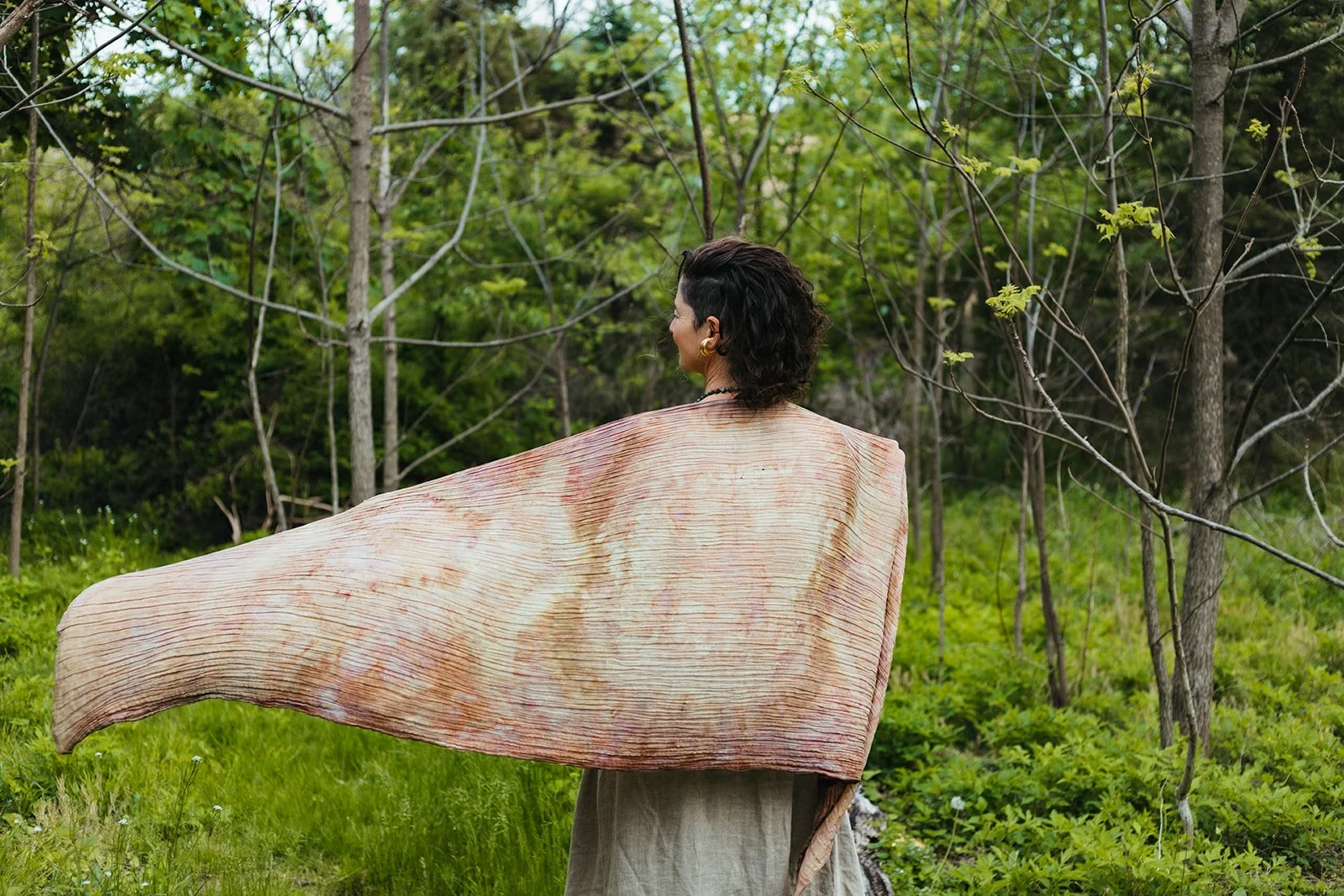 A woman with dark, wavy hair facing away, standing in a green outdoor setting with trees and grass, wearing a large, flowing beige and pink shawl.