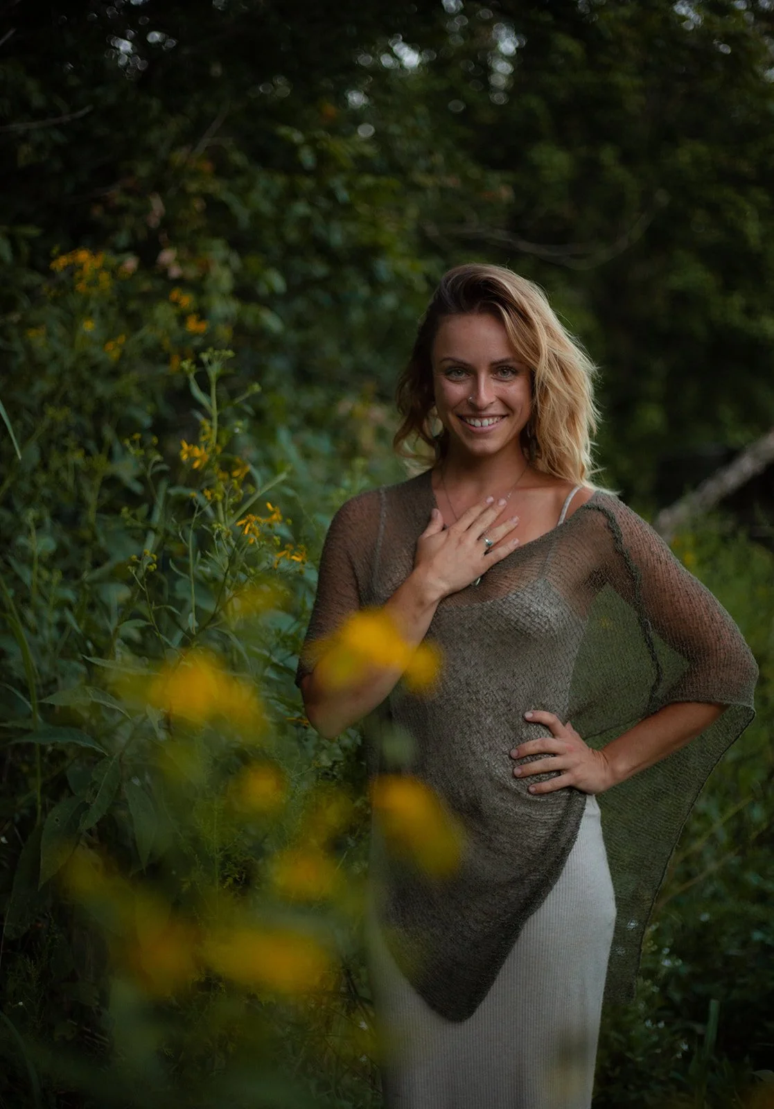 A woman with blonde hair smiling and standing outdoors among green foliage and yellow flowers. She is wearing a sheer, dark brown shawl over a light top and light-colored pants. Her right hand is on her chest, and her left hand is on her hip.