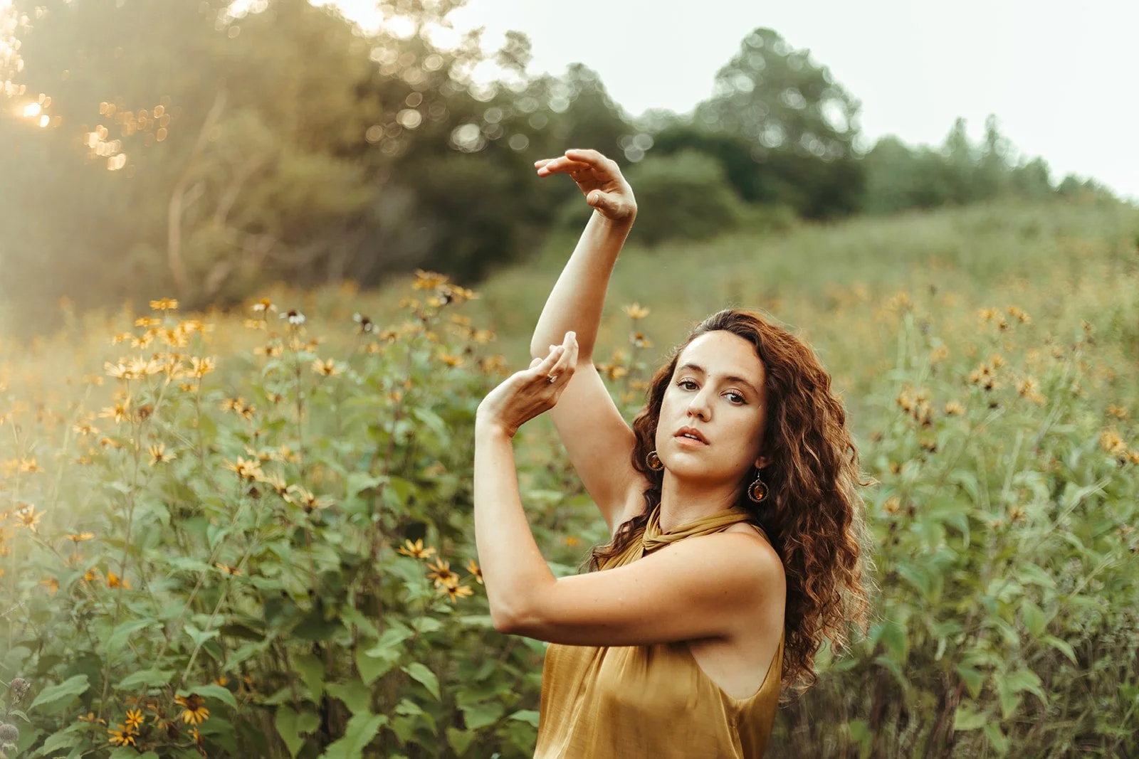 A woman with curly brown hair posing in a field of yellow flowers during daytime, wearing a sleeveless brown dress and earrings, with arms raised gracefully.