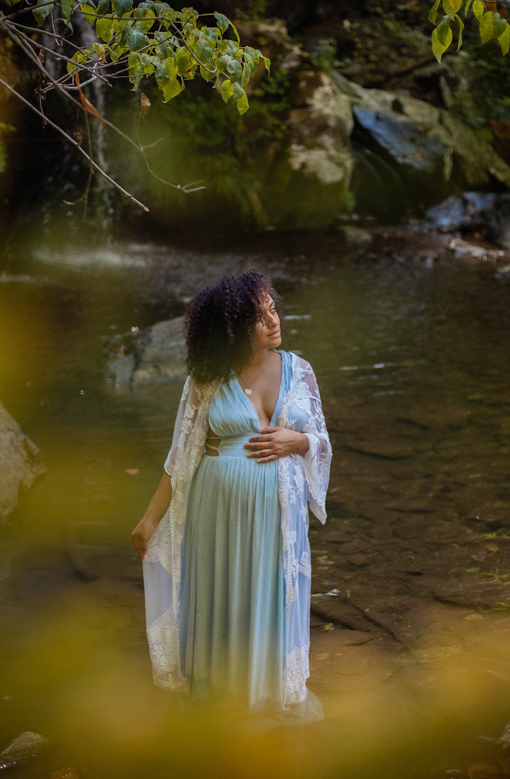 A woman standing in a shallow creek with rocks and greenery around, wearing a light blue flowing dress and a lace shawl.