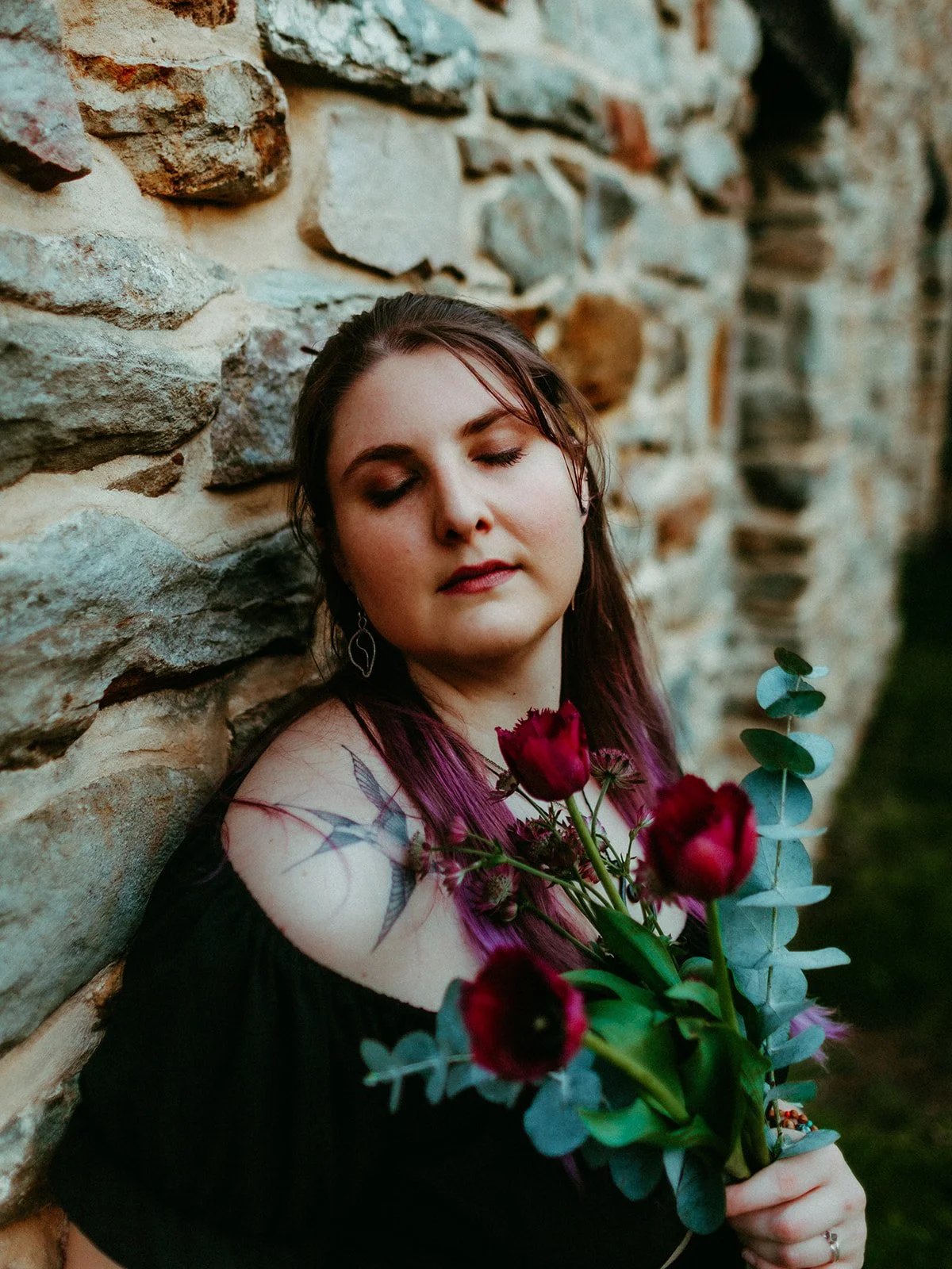 A woman with closed eyes and long dark hair with purple tips, holding a bouquet of red flowers and eucalyptus, leaning against an old stone wall.