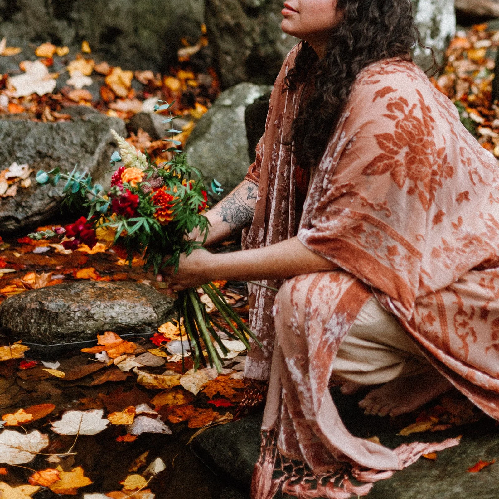 A woman kneeling by a creek in a forest, holding a colorful bouquet of flowers. She is dressed in a pink patterned shawl and beige pants with a tattoo visible on her forearm. Fallen autumn leaves surround her and float on the water.