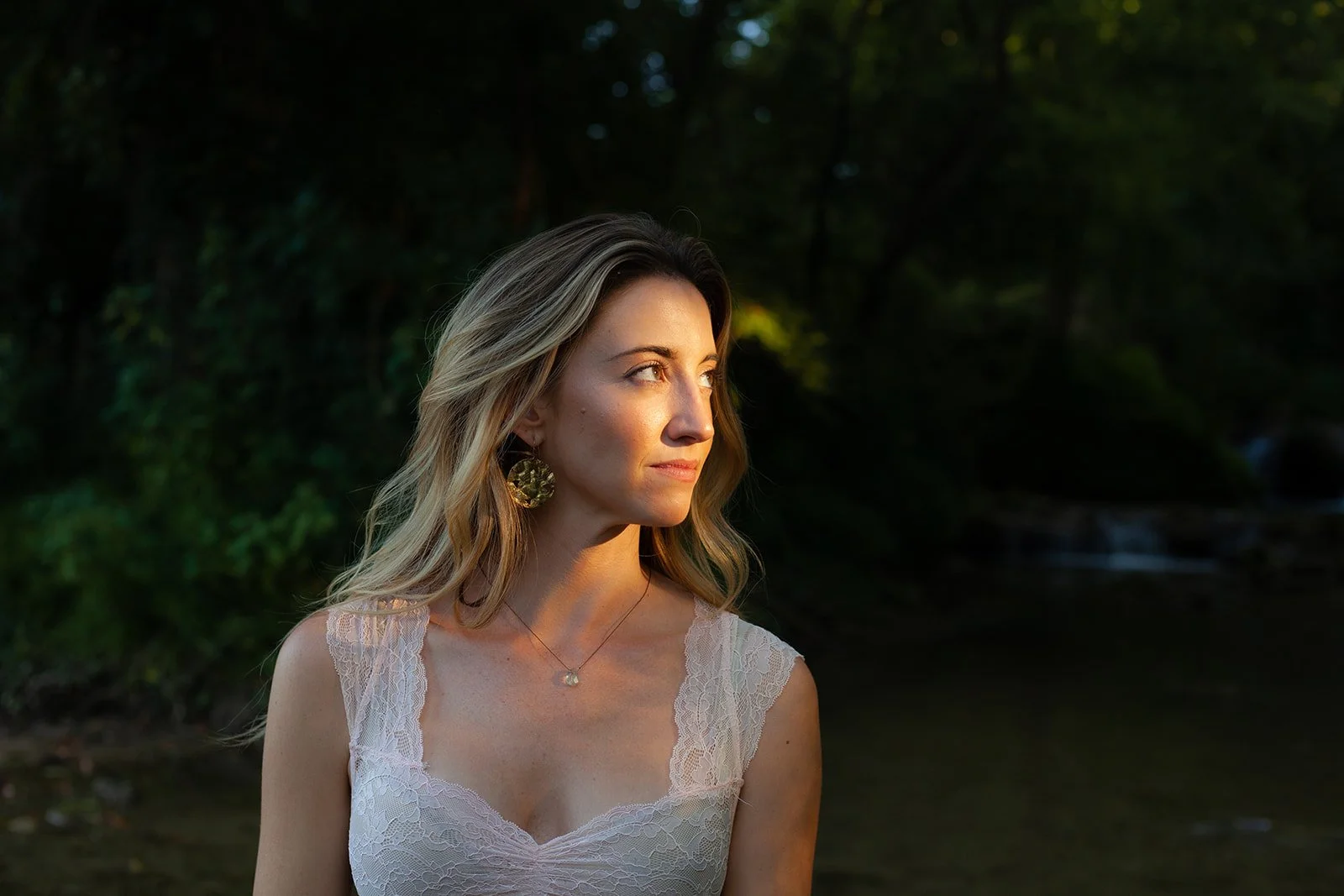 A young woman with wavy blonde hair looking to the side, wearing a lace white dress and gold earrings, standing outdoors in a lush green setting with sunlight illuminating her face.