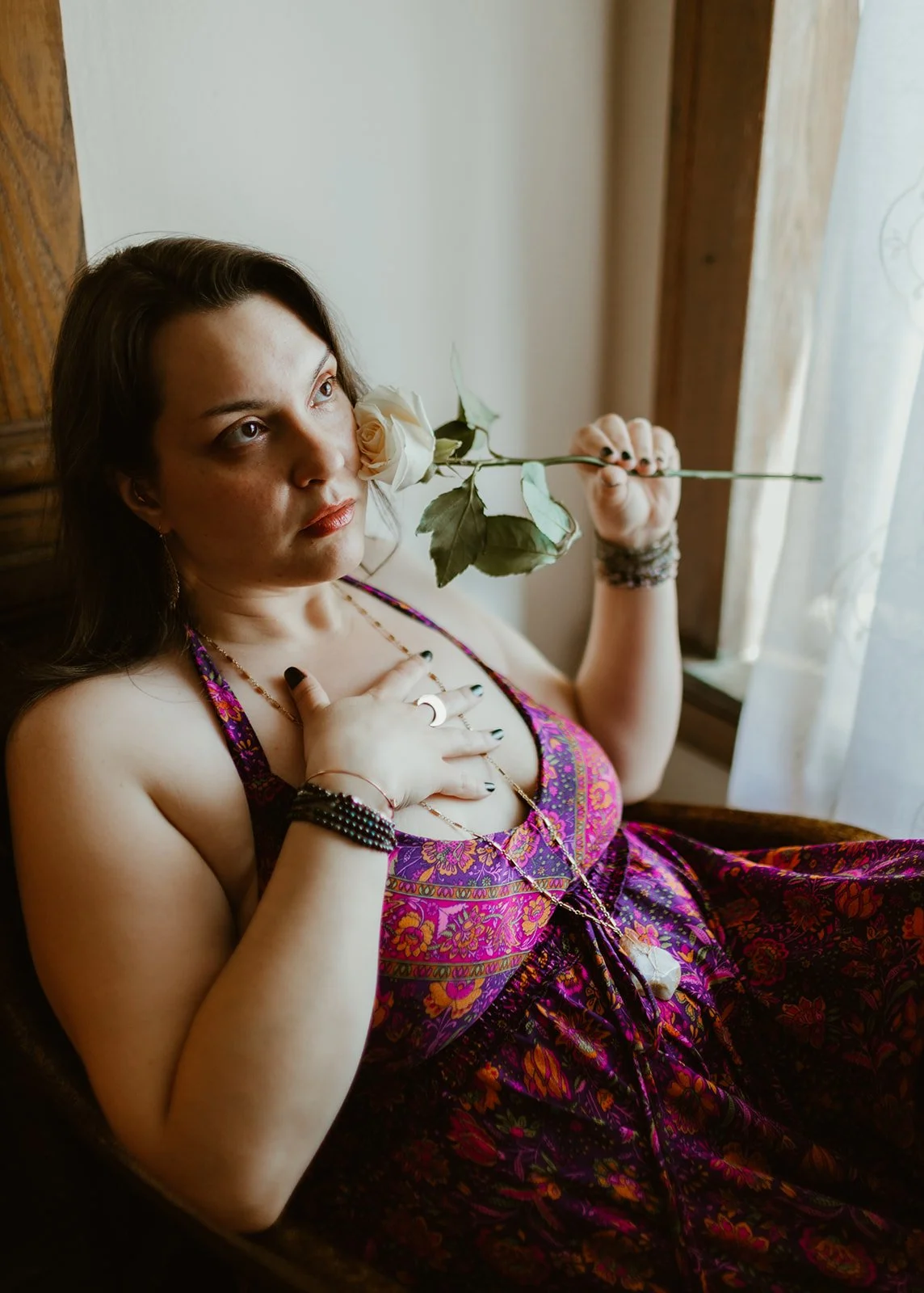 Woman with dark hair holding a white rose near her face, sitting by a window with curtains, wearing a colorful patterned dress, jewelry, and bracelets.