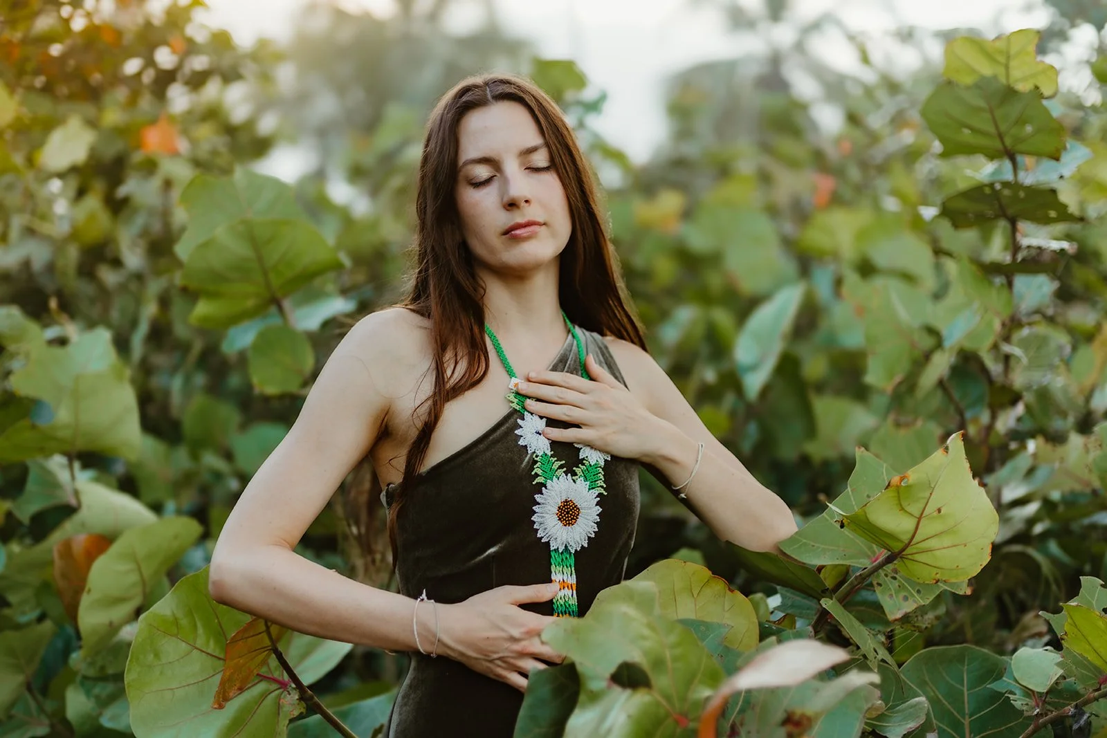 A woman with long brown hair and closed eyes standing among green leaves, wearing a black top and a beaded necklace with white daisy flowers.