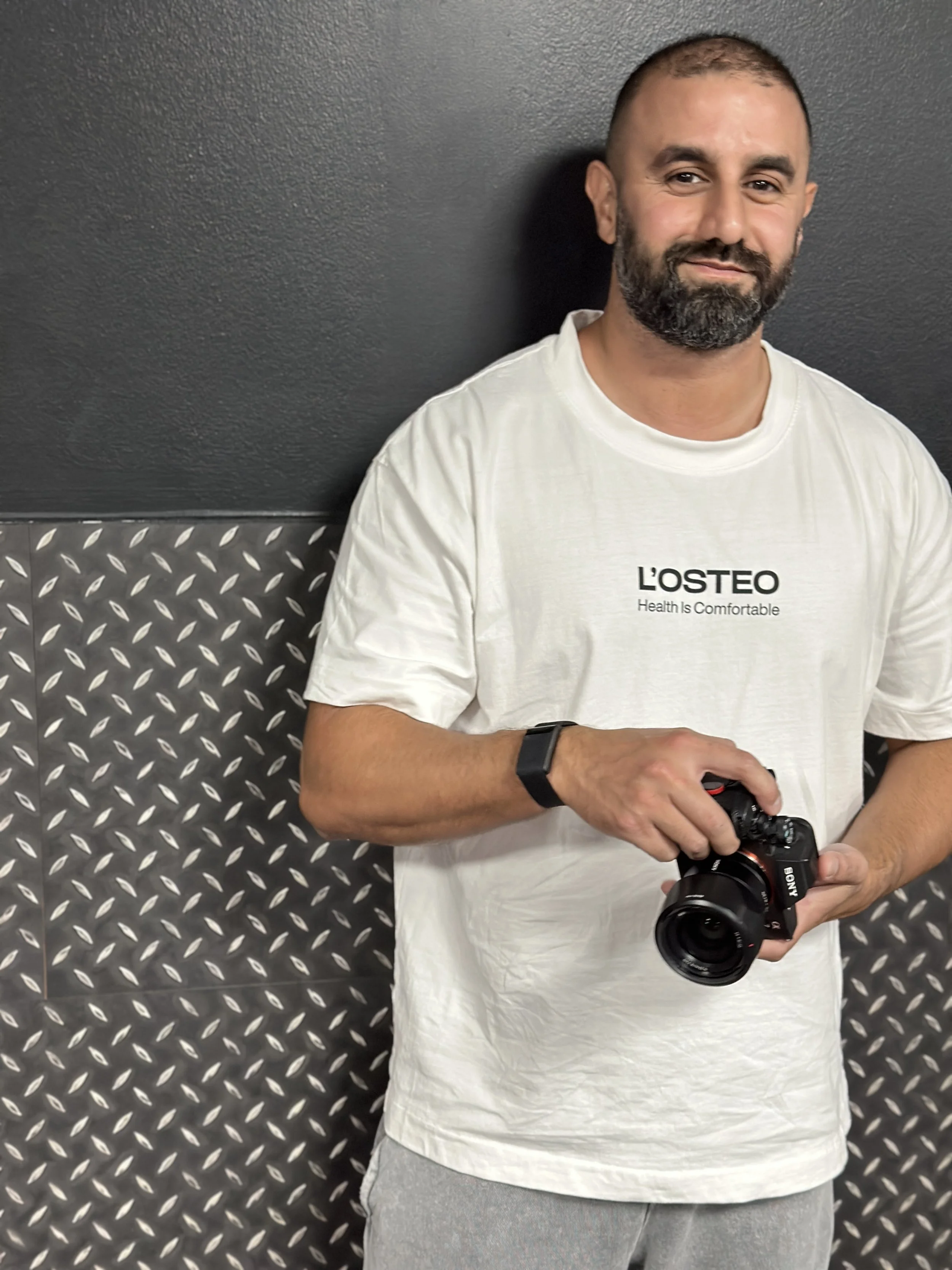 A man with a beard, wearing a white T-shirt and gray sweatpants, holding a camera in his right hand, standing against a dark wall with patterned metal paneling at the bottom.