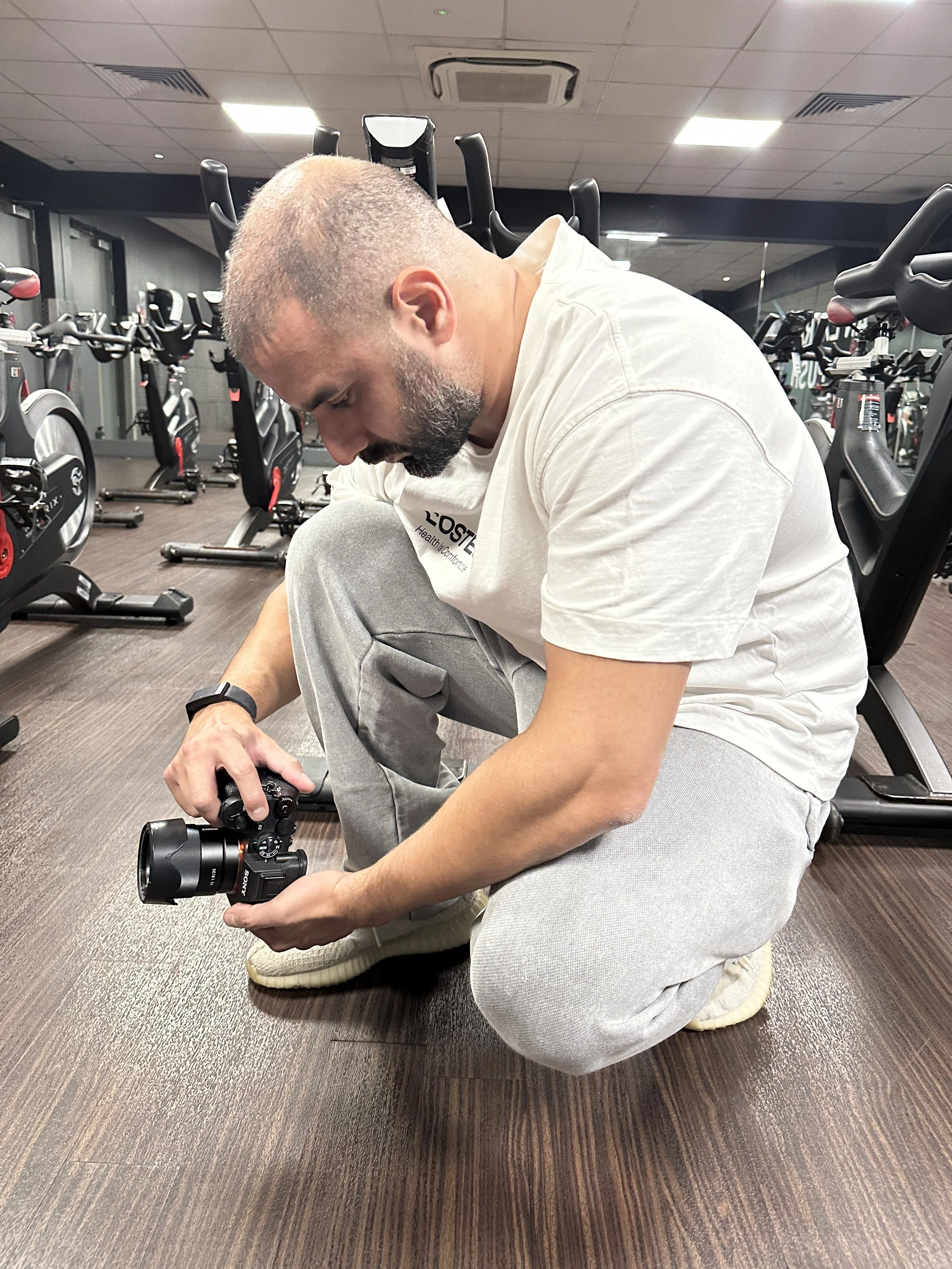 A man with a beard, wearing a white t-shirt and gray sweatpants, kneeling on a brown wooden floor in a gym, holding a Sony camera. The gym has multiple stationary bikes lined up against the wall, with a black ceiling and fluorescent lighting.