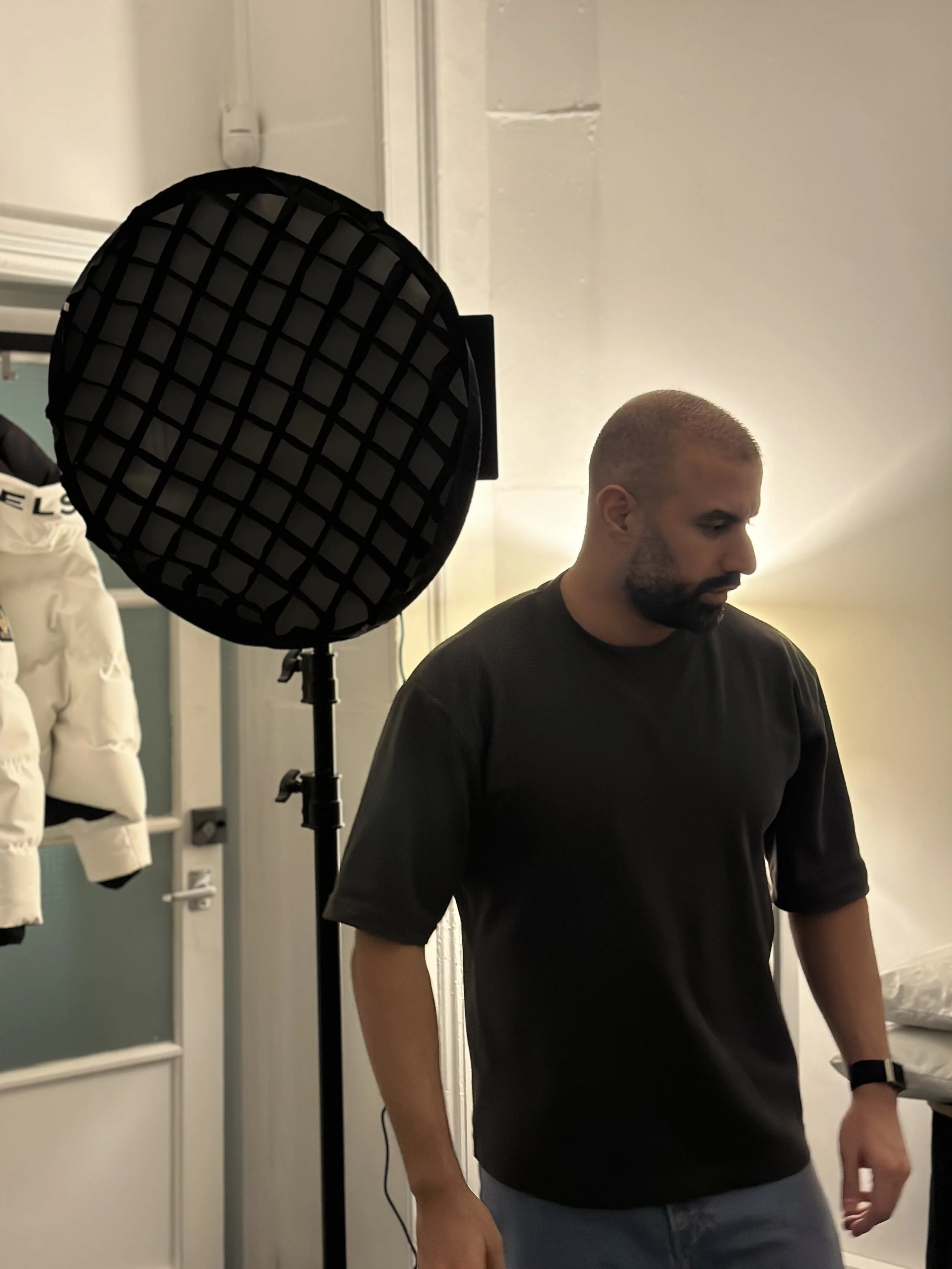 A man with short hair and a beard wearing a black shirt, standing indoors near a door, with a soft light illuminating his face. There is a black studio light with a grid filter on a stand nearby, and a white appliance is visible in the background.